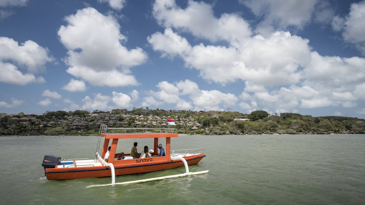 An outrigger canoe glides along calm, green water as four passengers look toward a distant shore dotted with trees and huts.