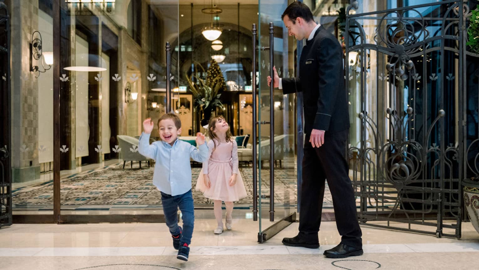 A young boy and girl walking through a hotel door being held open by a man in a suit.