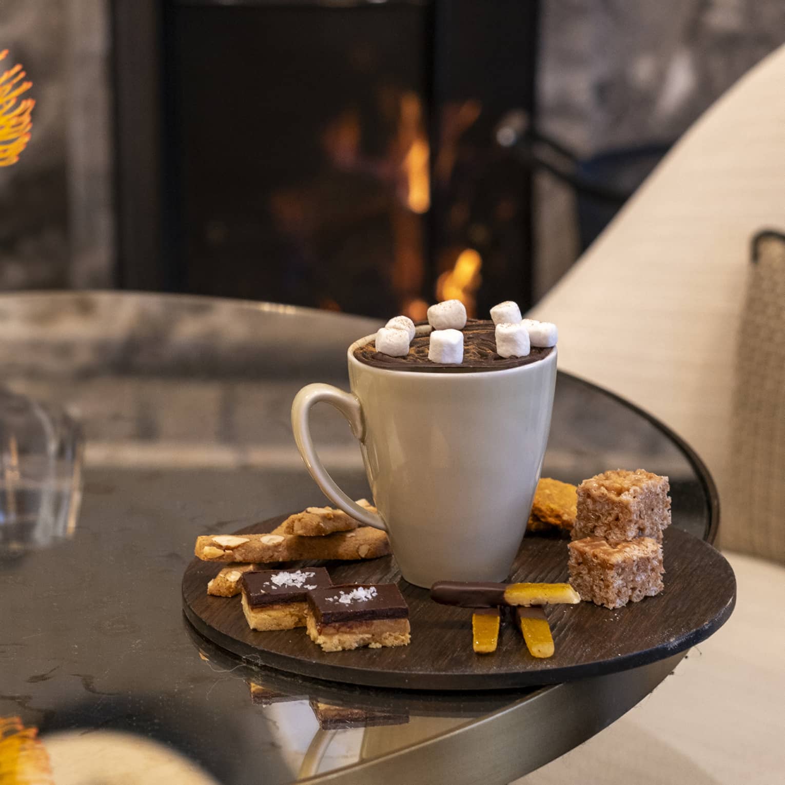 White mug of hot chocolate with mashmallows on a dark, round platter filled with small desserts