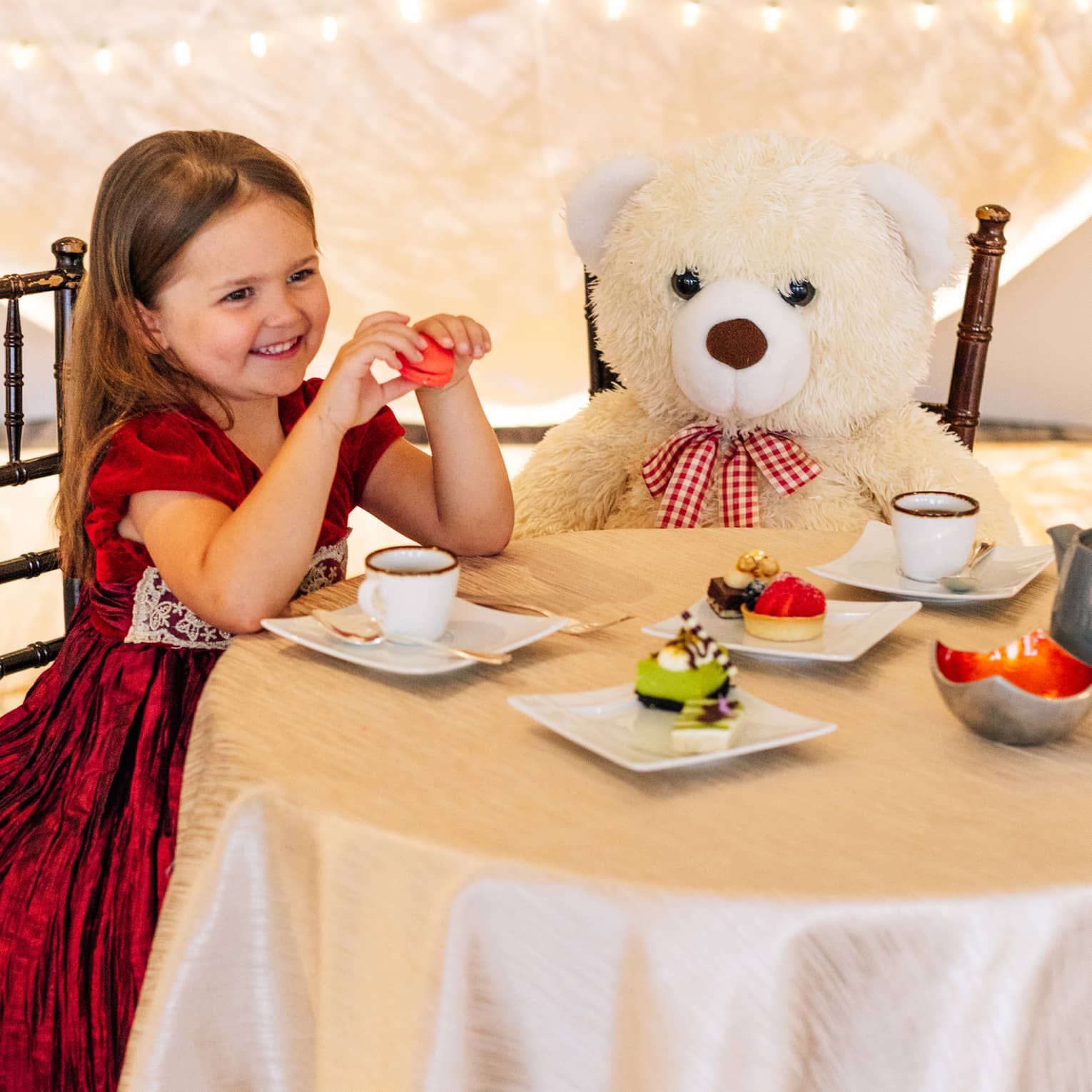 A small girl in a red dress sitting with a large teddy bear at a table with tea and small desserts.