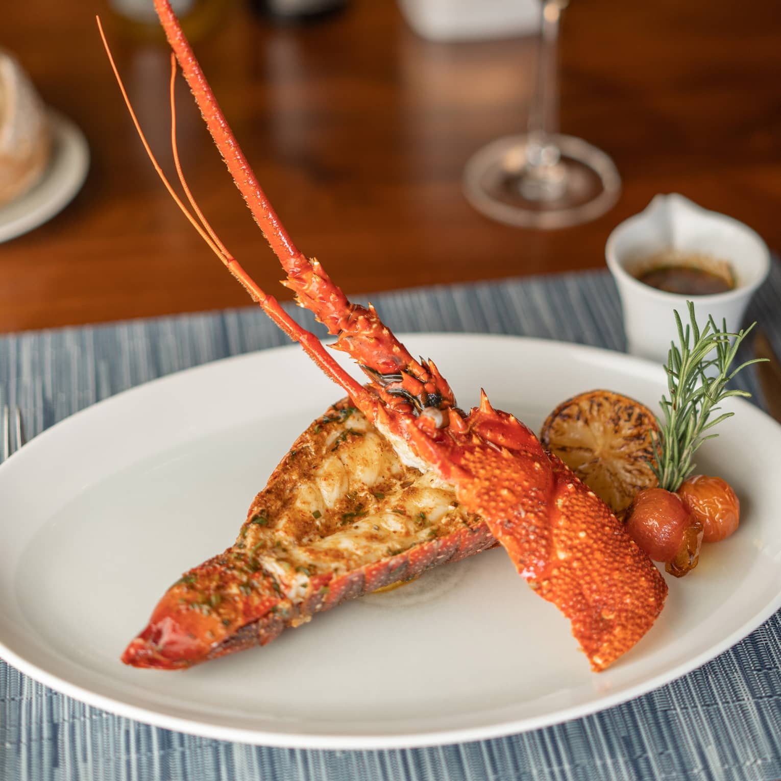 Halved rock lobster artfully arranged on plate with charred half lemon, rosemary sprig and roasted cherry tomato garnish on white plate, glass of white wine in background