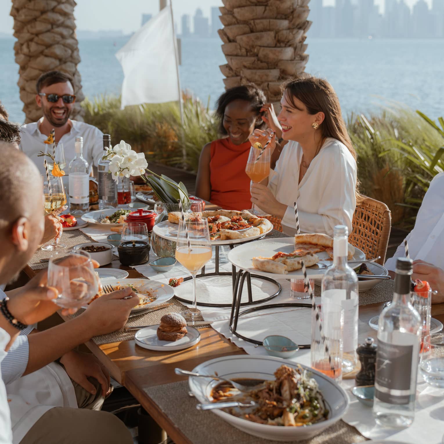 Group sits around long outdoor dining table eating brunch with the ocean and a city skyline in the background