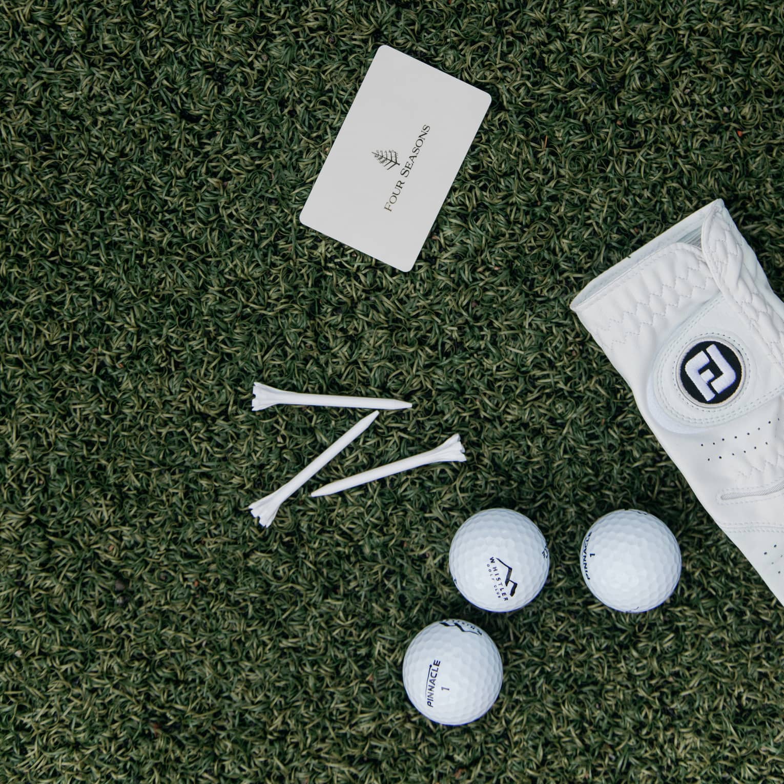 On green turf, a Four Seasons card, three golf balls, three tees and a white leather golf glove dotted with tiny holes.