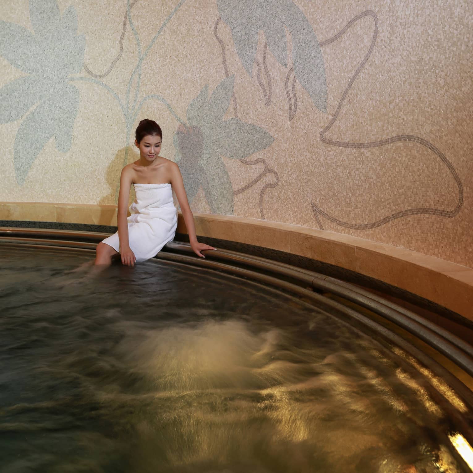 Woman wearing white towel sits on edge of round tile whirlpool in spa