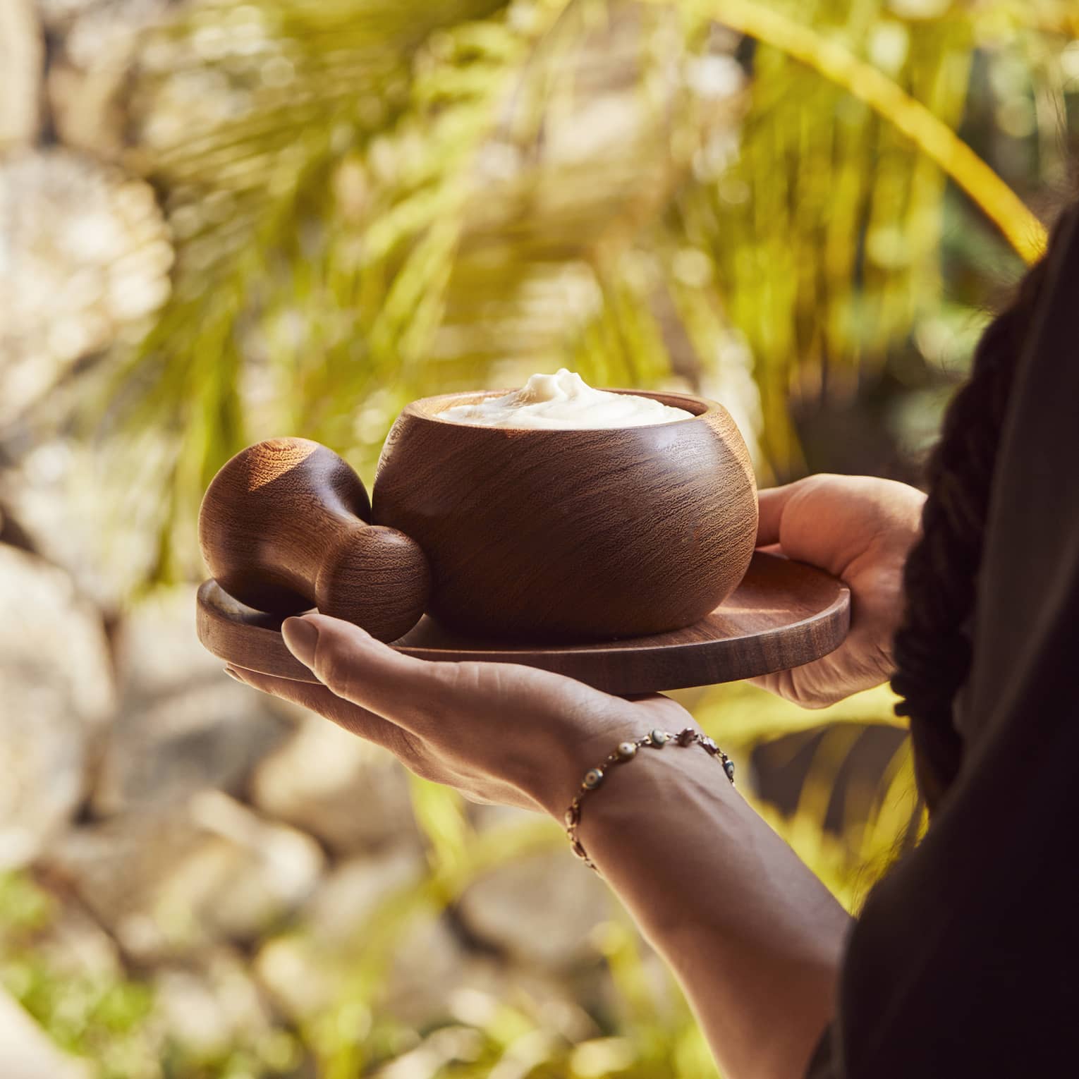 Two hands carry a small round tray with a wooden pestle and a bowl filled with a creamy body scrub past golden palm fronds.
