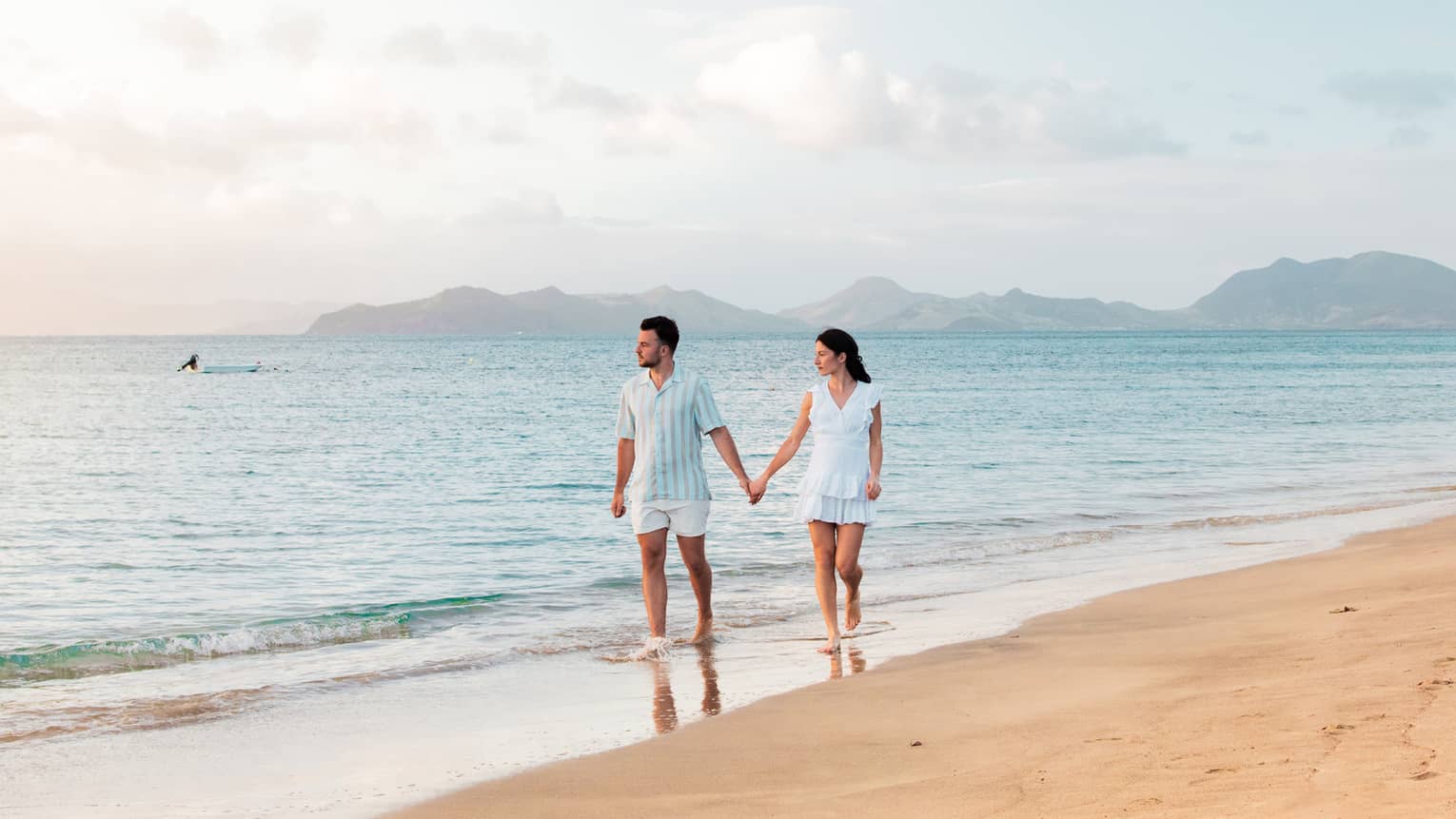 Two guests wade hand in hand at the edge of a sandy beach, white clouds dotting the sky and low mountains visible beyond.