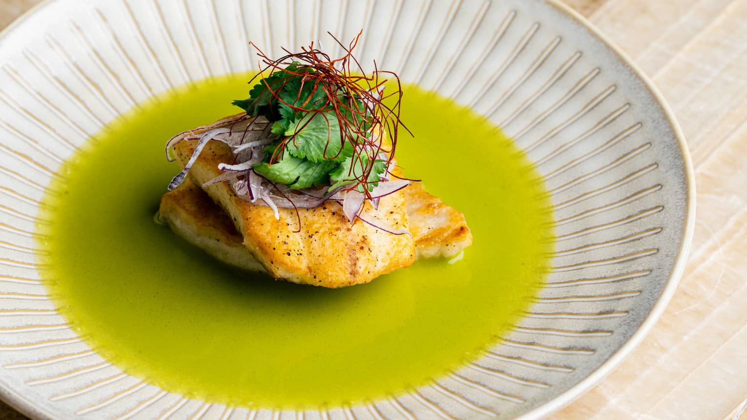 Golden-brown pieces of fish sit on vibrant green sauce in a pottery bowl beneath a generous garnish of cilantro and saffron.