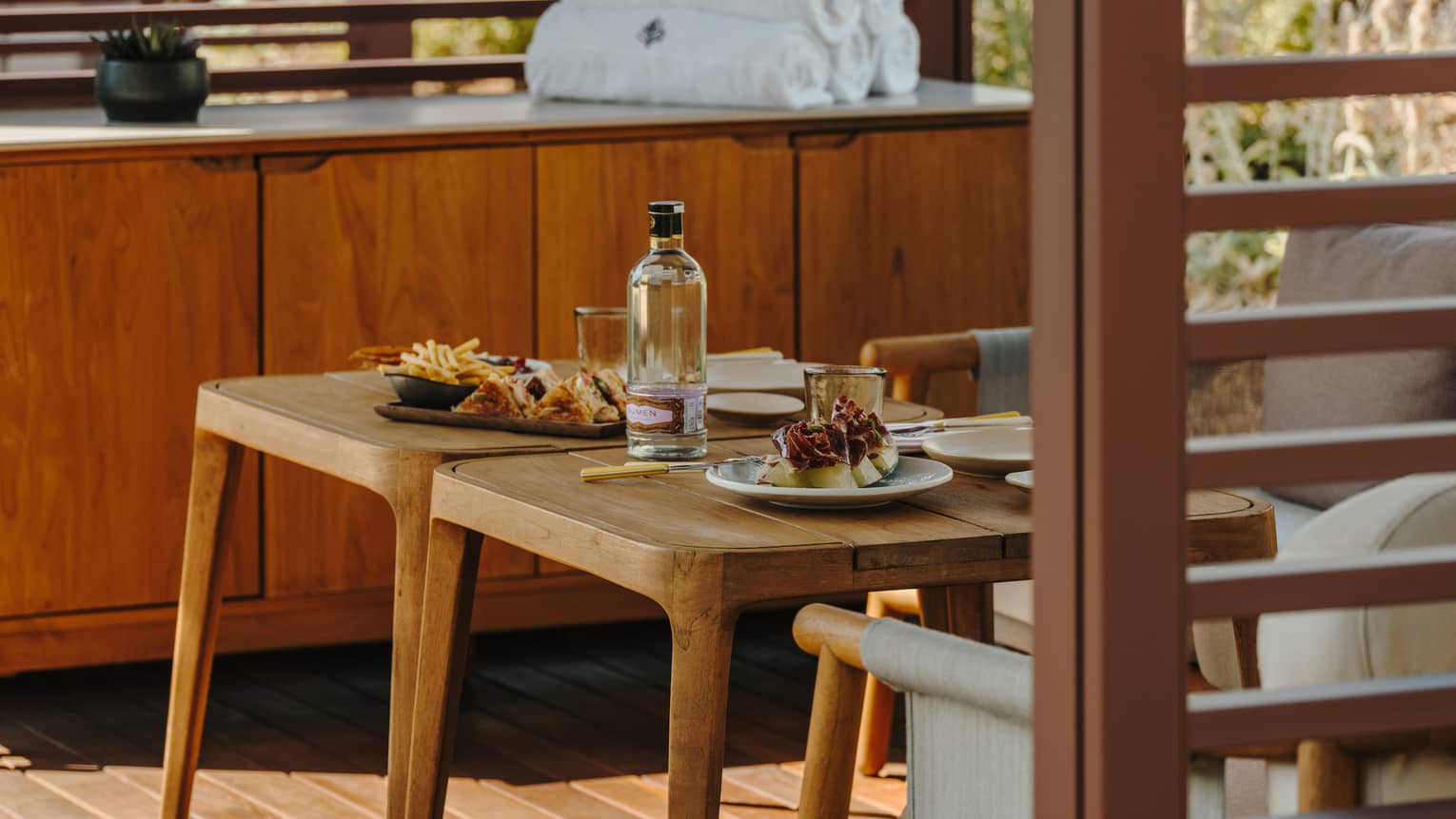 A private pool cabana with small tables, comfortable seating, a cabinet with rolled-up towels and slatted walls admitting filtered light sunlight.