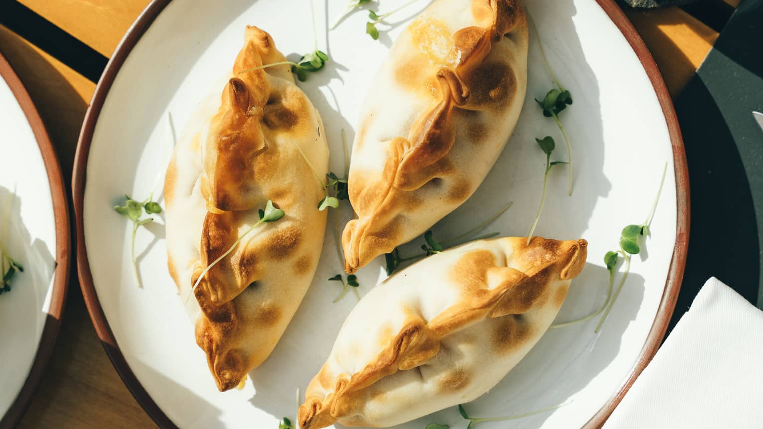 Three plump, golden-brown empanadas on a porcelain dish surrounded by sunlit water glasses and other plates of fine foods. 