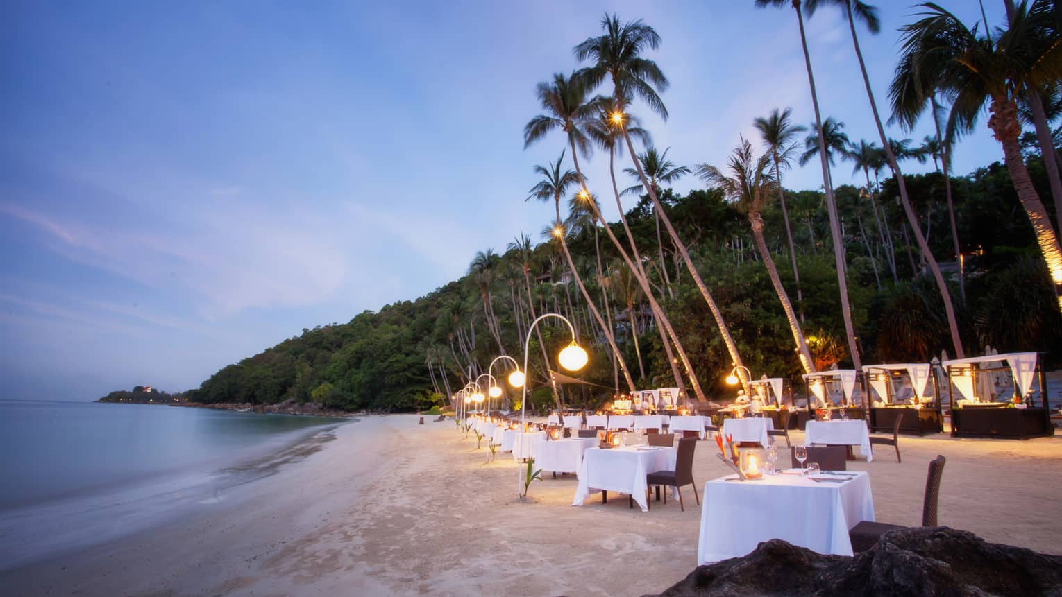 Dining tables set up along white sand beach under palm trees at dusk