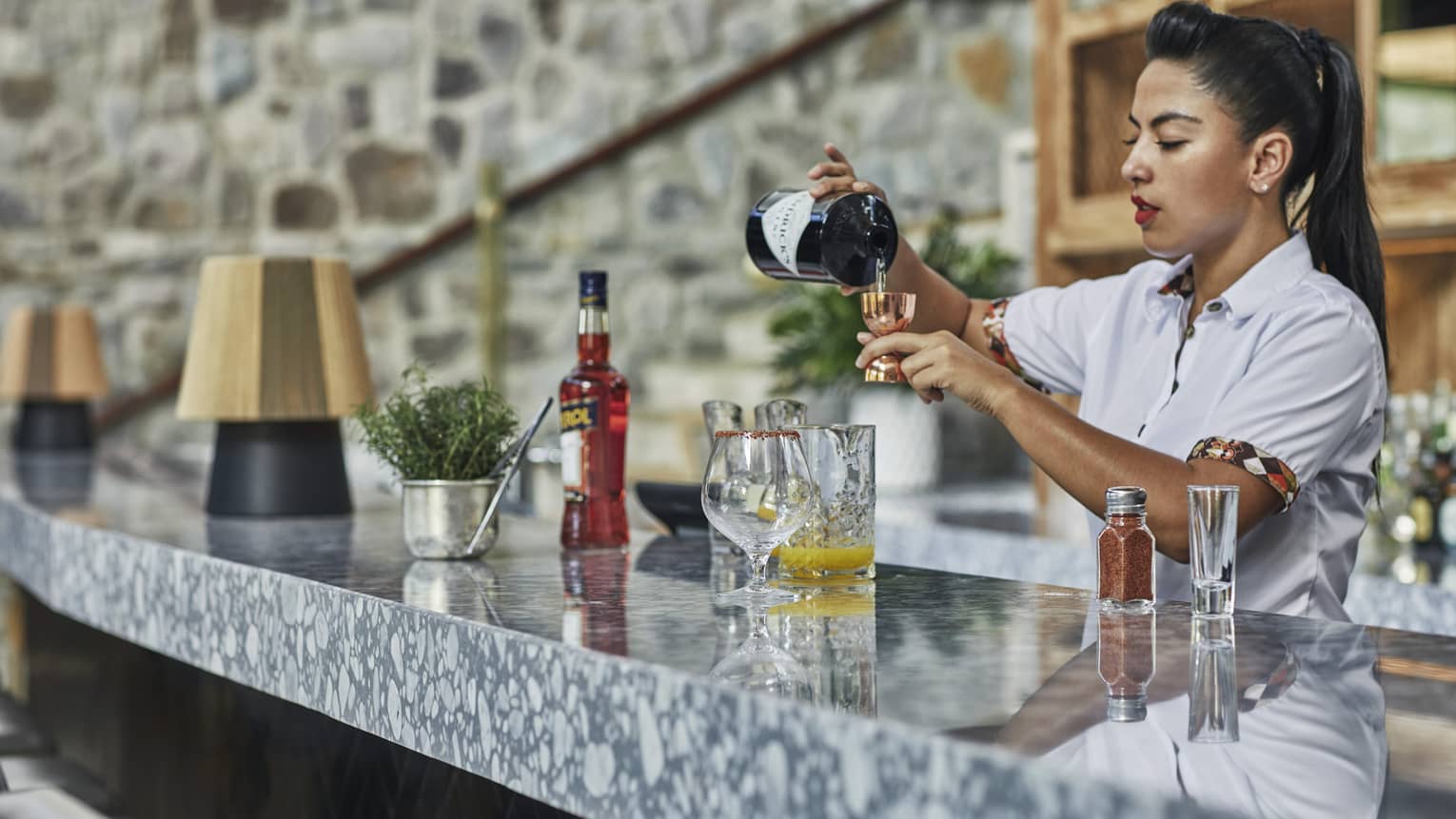 A woman with a high ponytail prepares a drink behind a marble countertop.