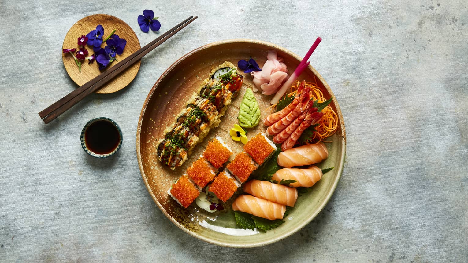 A ceramic bowl holding sushi and sashimi rolls, a small container of soy sauce, a small ceramic plate holding chopsticks and small purple flowers