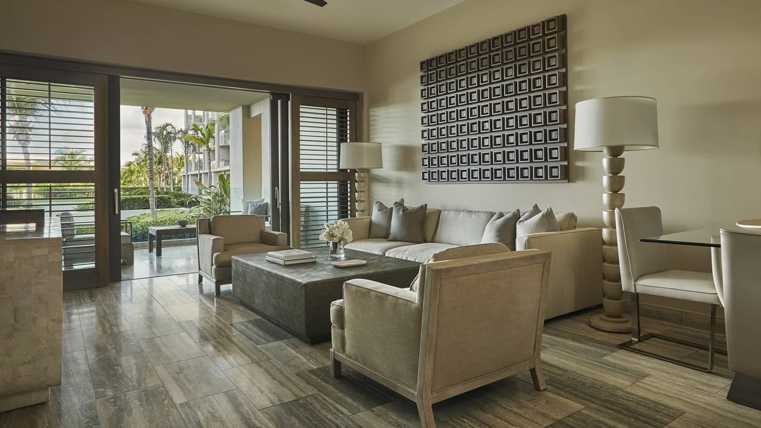 Cozy living room with a beige sofa, armchairs, a modern coffee table, decorative wall art and sliding doors leading to an outdoor patio with tropical views
