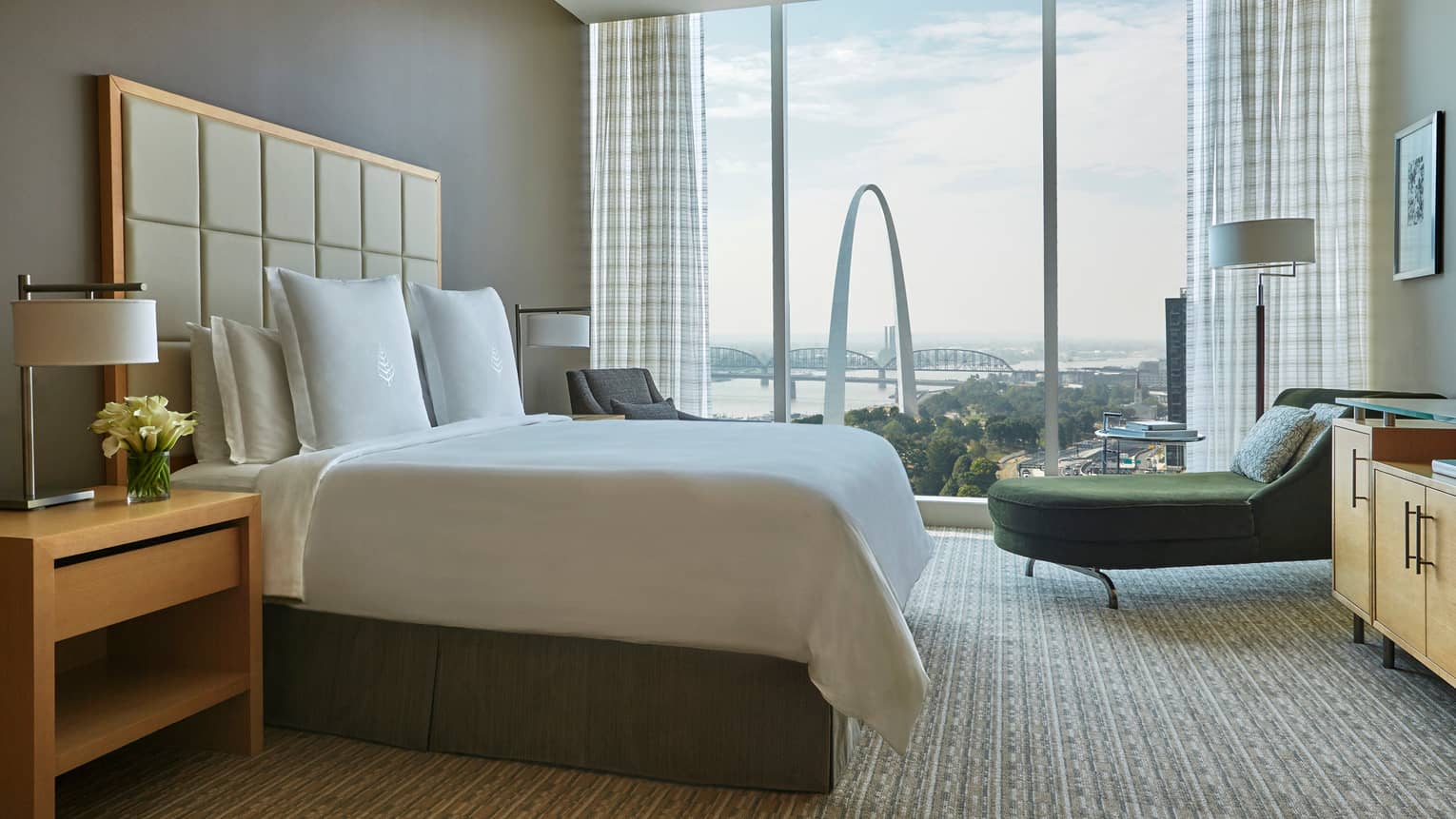 Bed with tall, padded white headboard, green velvet chaise beside floor-to-ceiling window with arch view