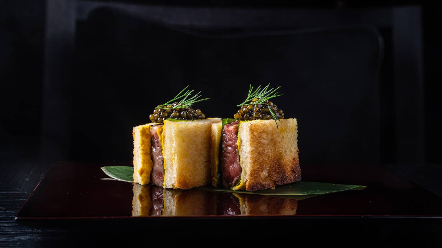 Sando Wagyu Beef topped with Caviar on banana leaf, dill frond garnish, black background