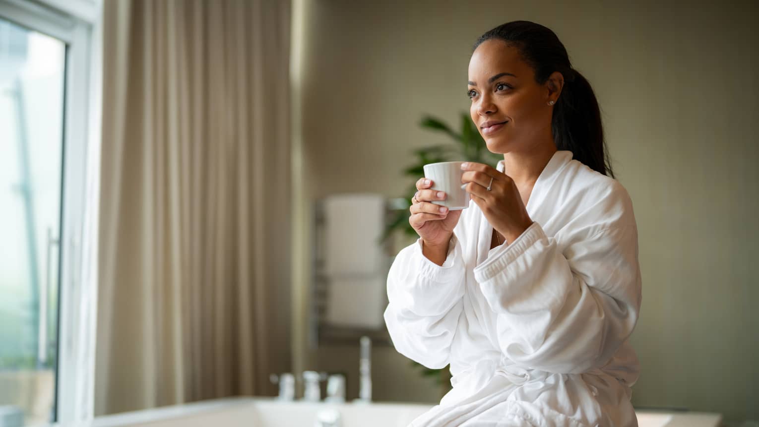 A woman dressed in a white bathrobe sits on the edge of a large bathtub while holding a beverage in a white ceramic mug