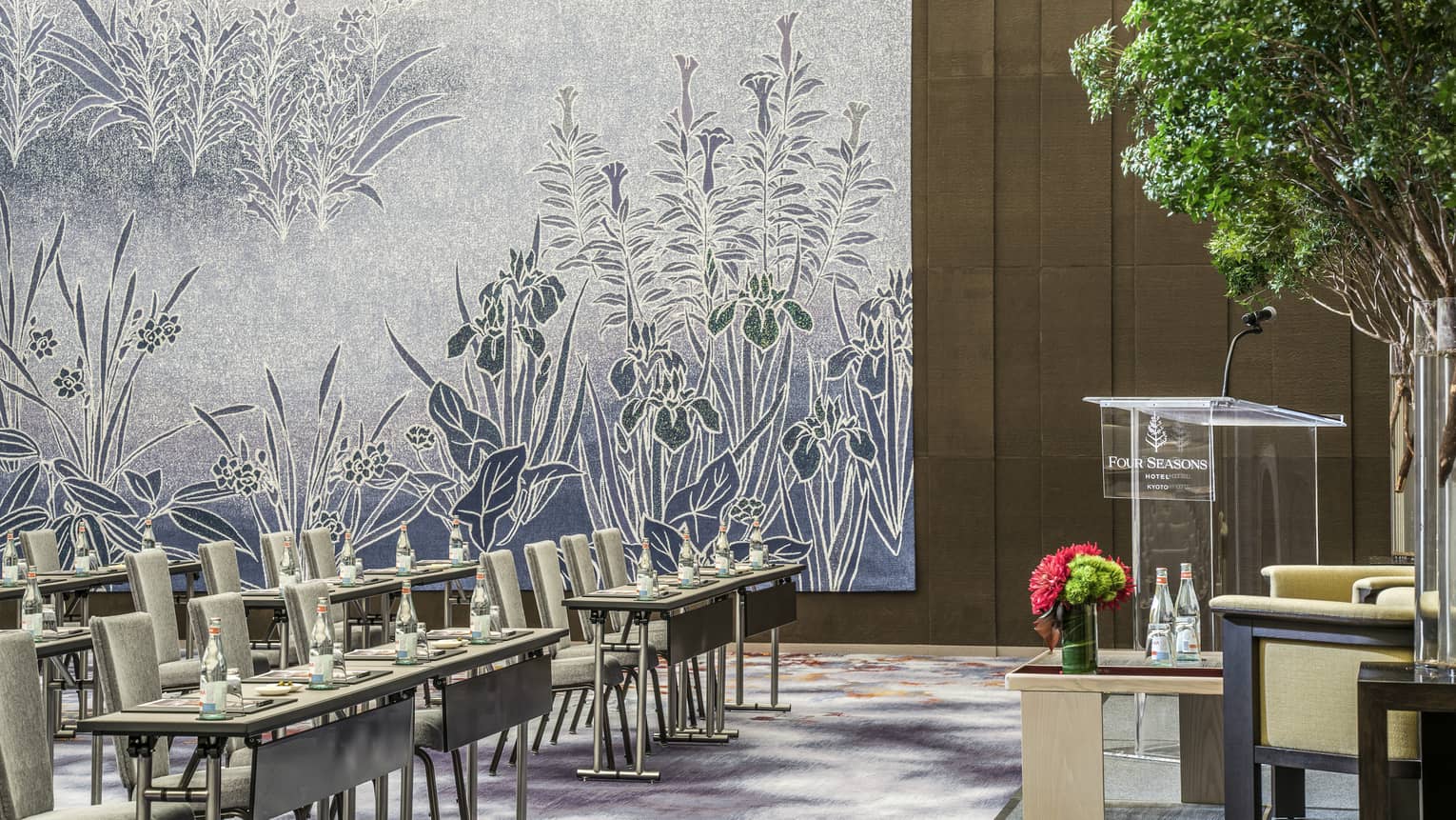 Conference in ballroom, rows of meeting tables, chairs along floral accent wall, facing podium