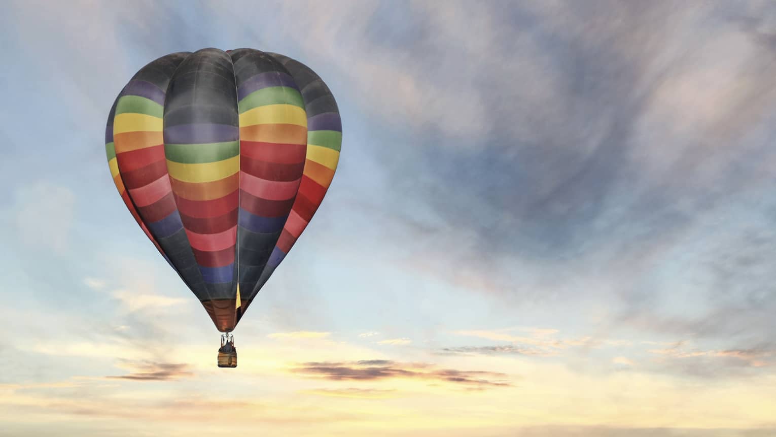 Rainbow-striped hot air balloon floats in the sky at sunset