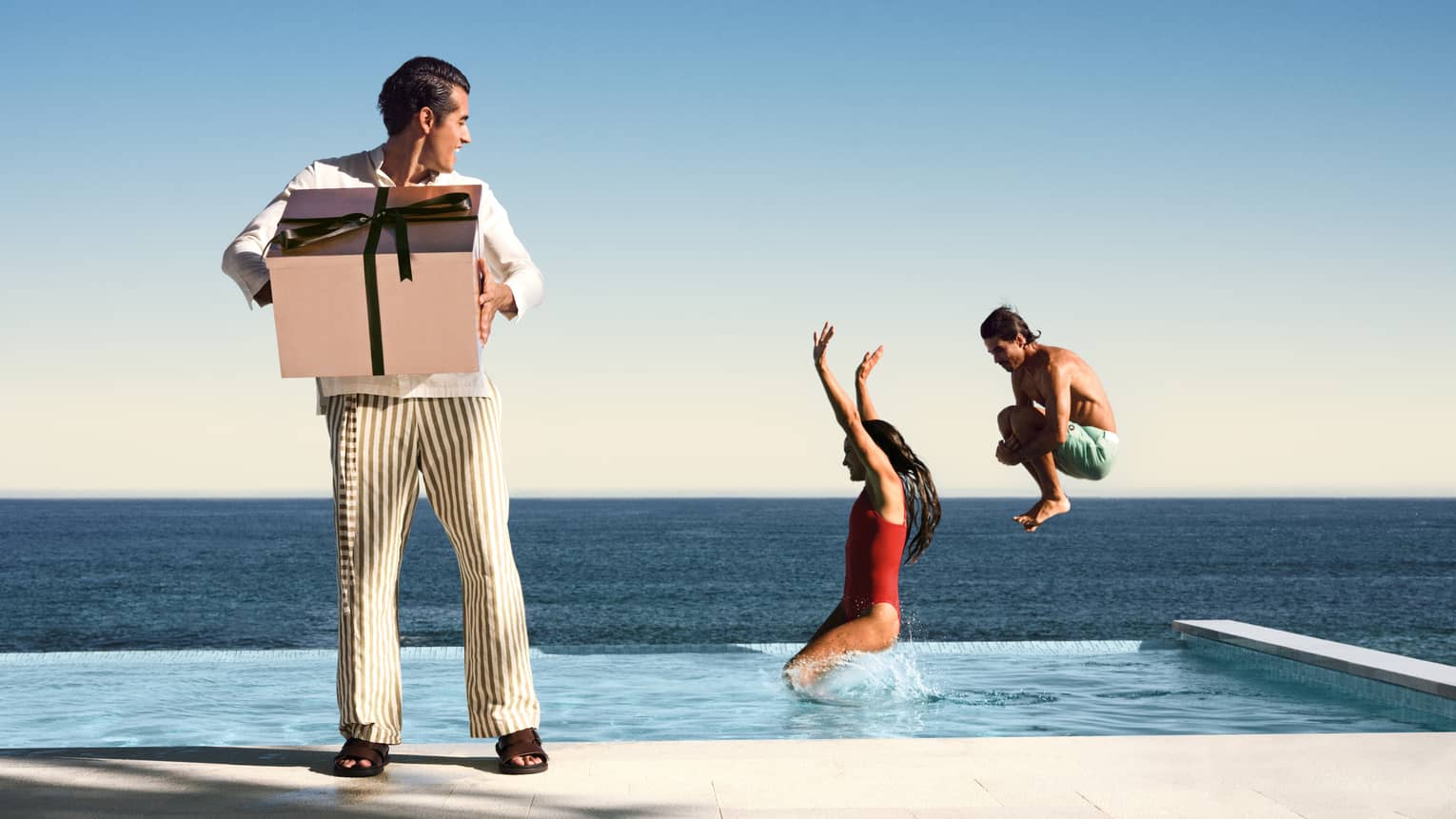 Two people jump into an infinity pool with ocean view, while a man holds a large present