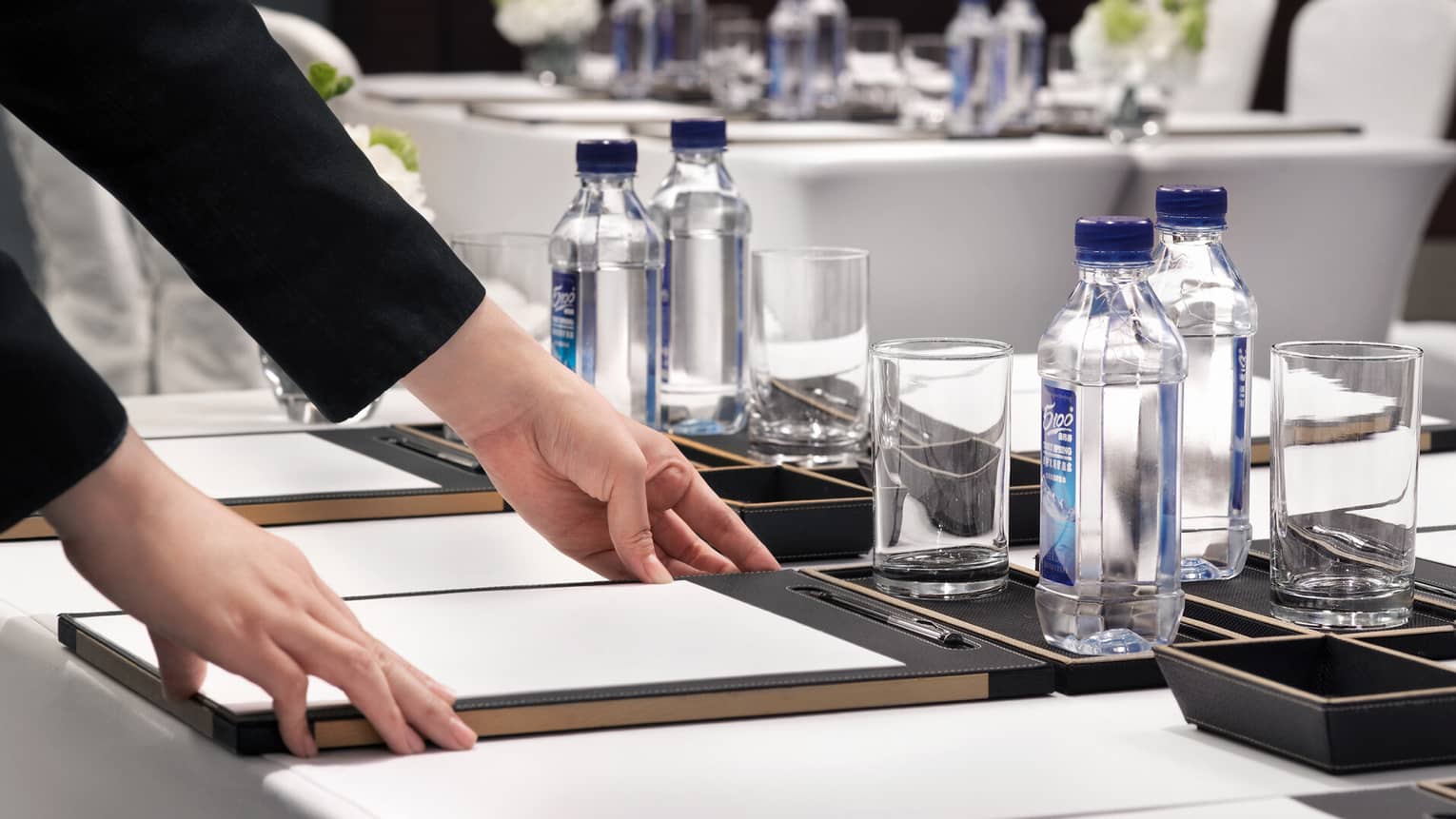 A person adjusts a writing pad on a conference room table with bottled water, glasses and floral arrangements
