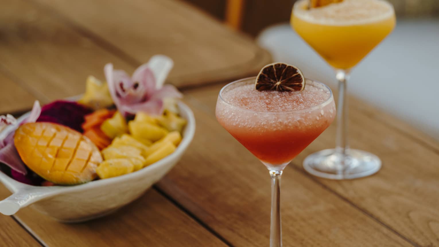A wood table holds two stemmed glasses of pink and orange drinks with garnishes and a bowl of small-bite food items.