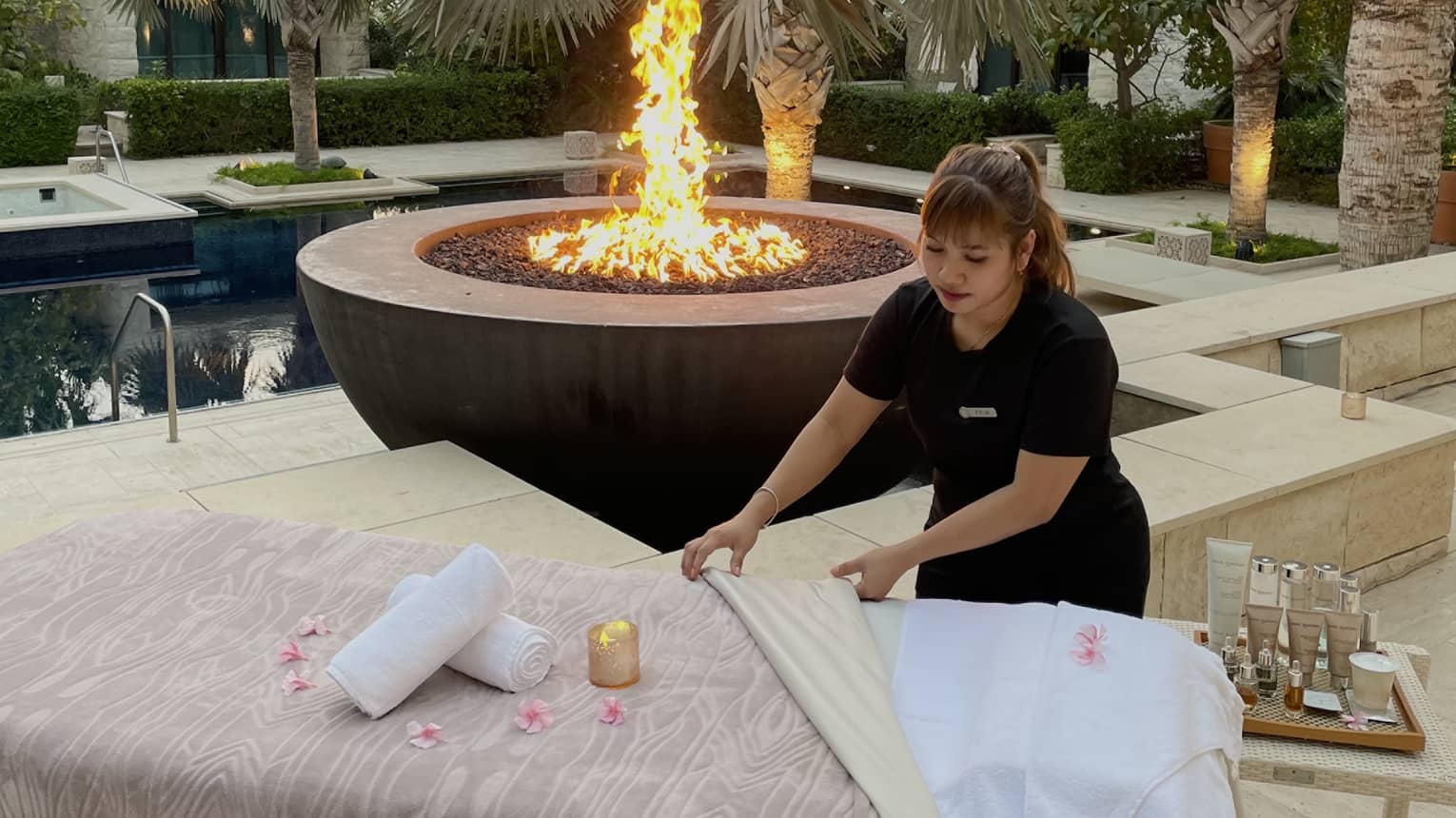 Spa attendant sets up a massage table next to a large firepit by an outdoor pool surrounded by palm trees