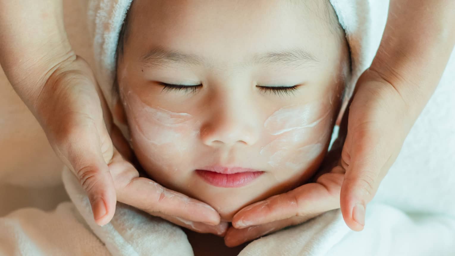 A close-up of a relaxed child wrapped in a towel and robe and enjoying a masseuse massaging a cream into their face.