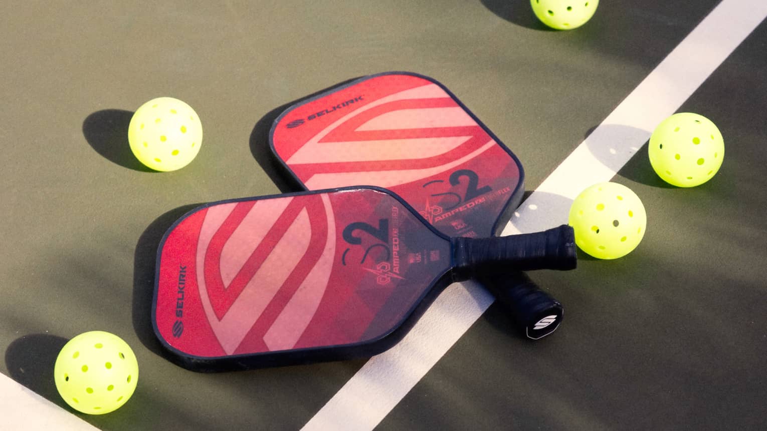 Close-up of two red and pink pickleball racquets, handles crisscrossed on a white court line, surrounded by five pickleballs.