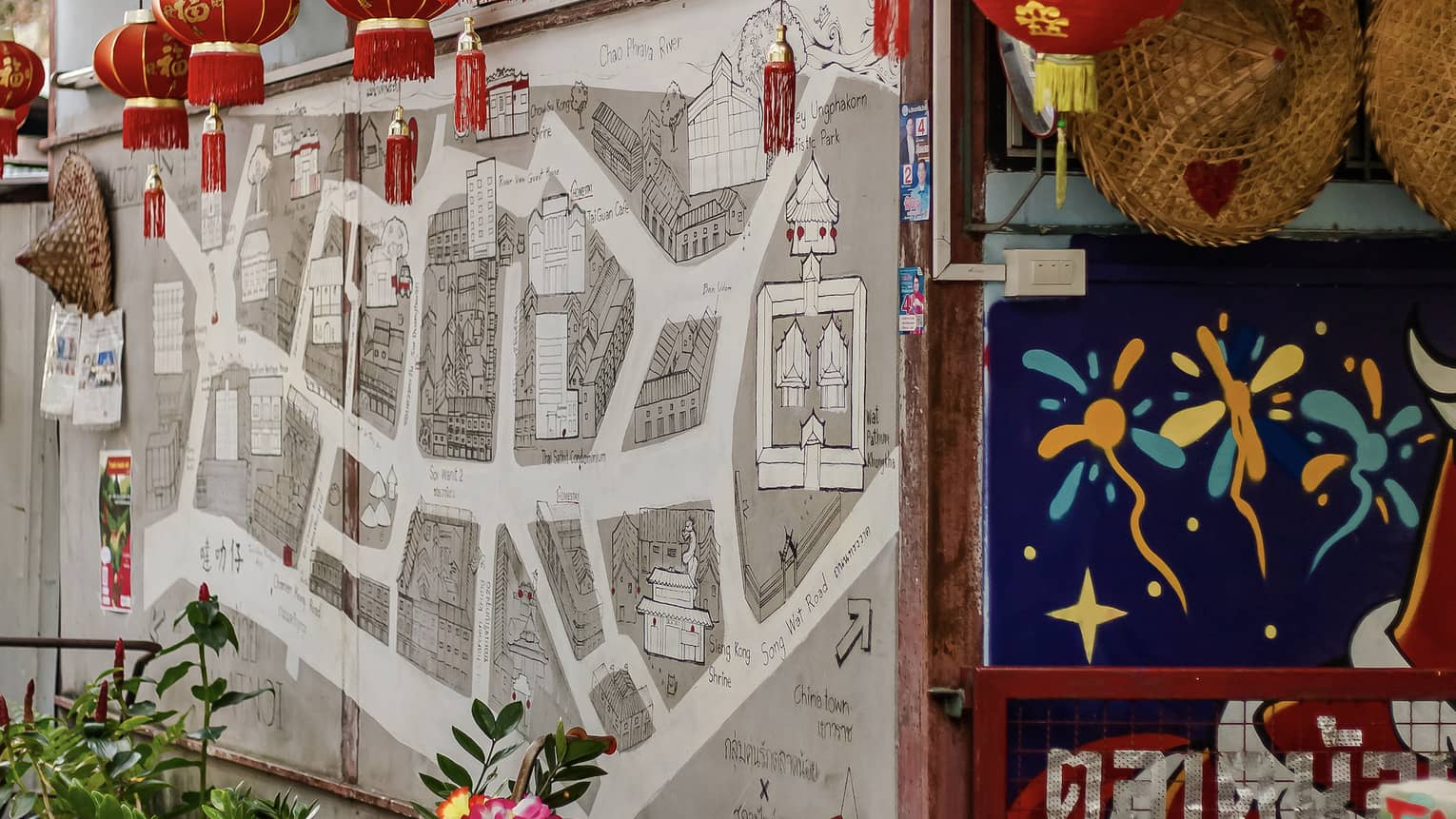 Red and gold paper lanterns hang from roof over colourful wall murals, doorway, potted plants