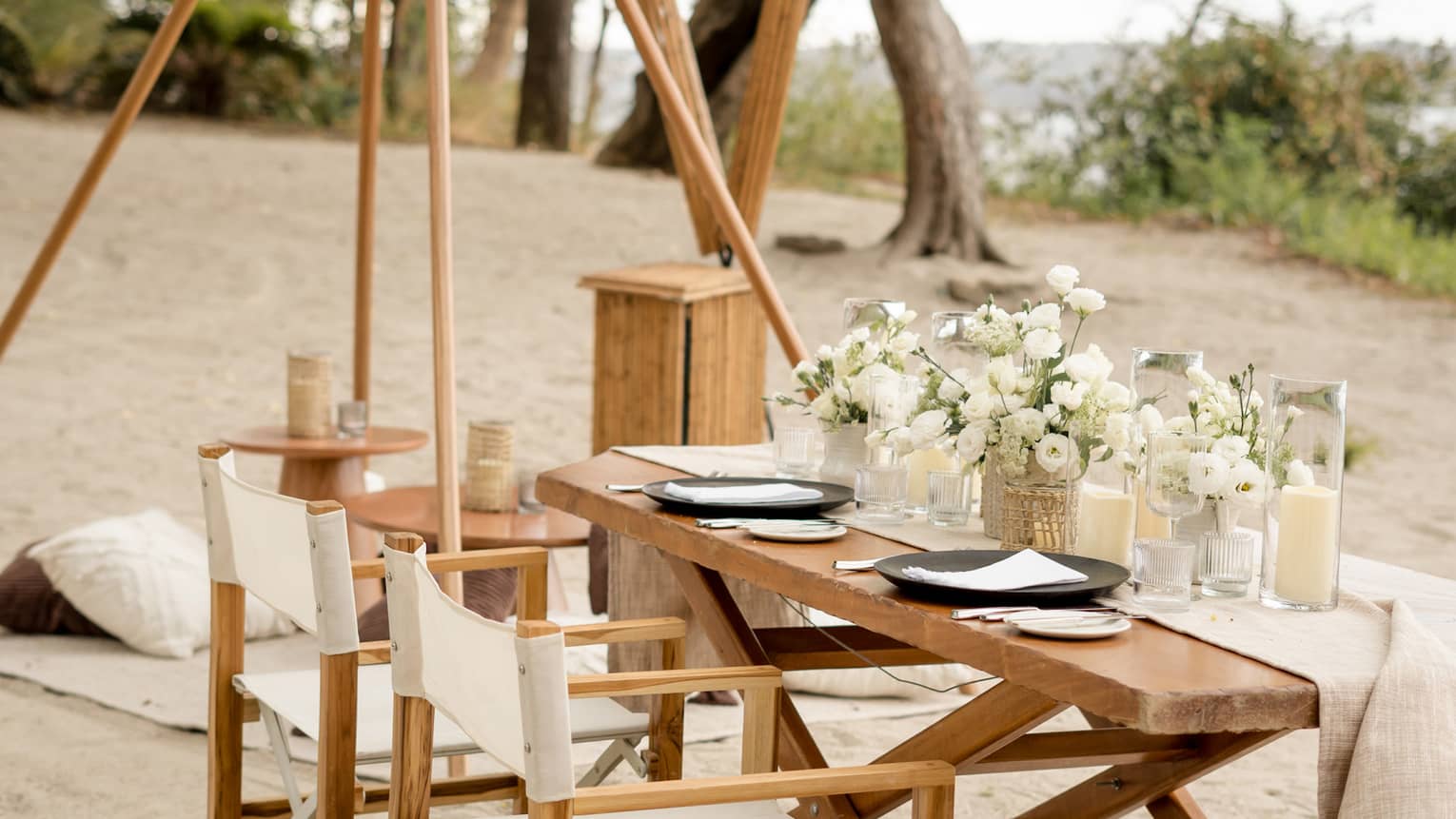 Wooden dining table and chairs set up on a beach, decorated with white floral centrepieces and candles