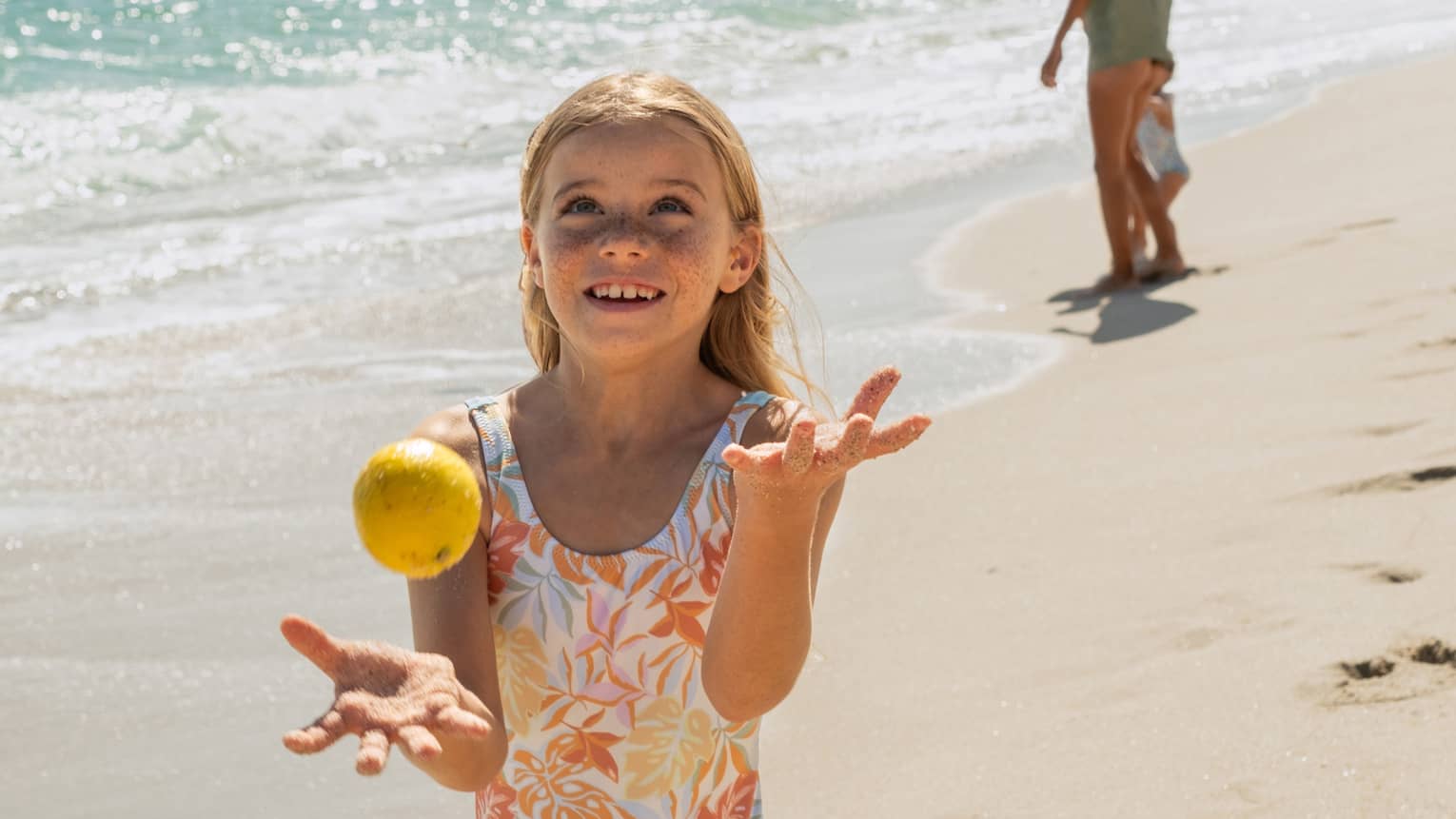Child juggling lemons on a sunny beach with gentle ocean waves in the background, while another person walks along the shoreline in the distance.