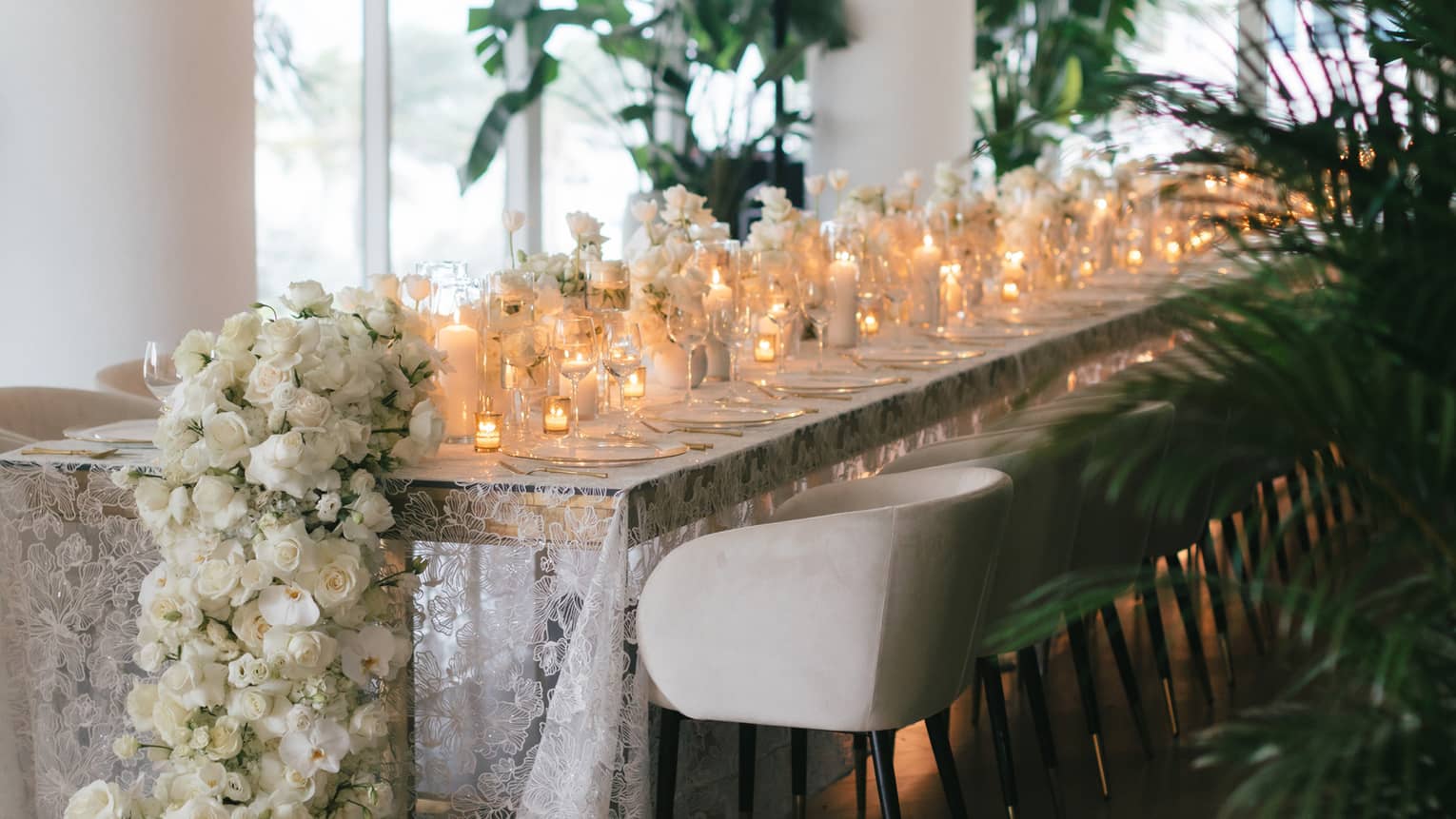 Long rectangular table topped with a white lace tablecloth and a variety of votive candles and white floral garlands lining the centre
