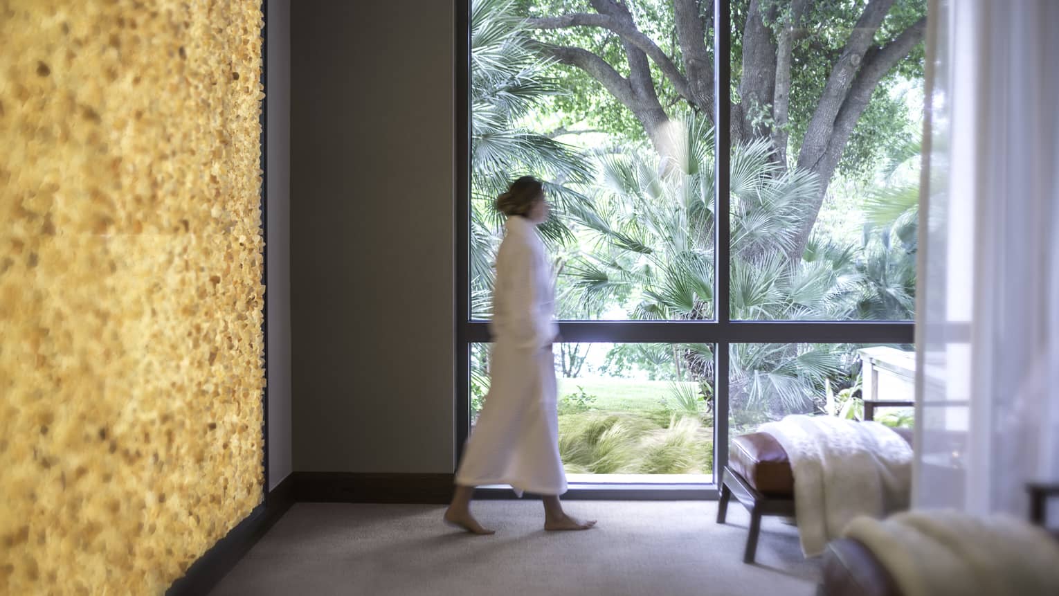 Woman wearing white robe walks down spa hall past sunny window