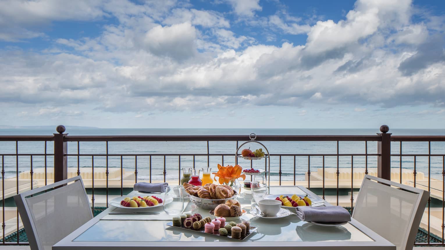 Balcony table set with fresh pastries, cakes, fruits, sea in background