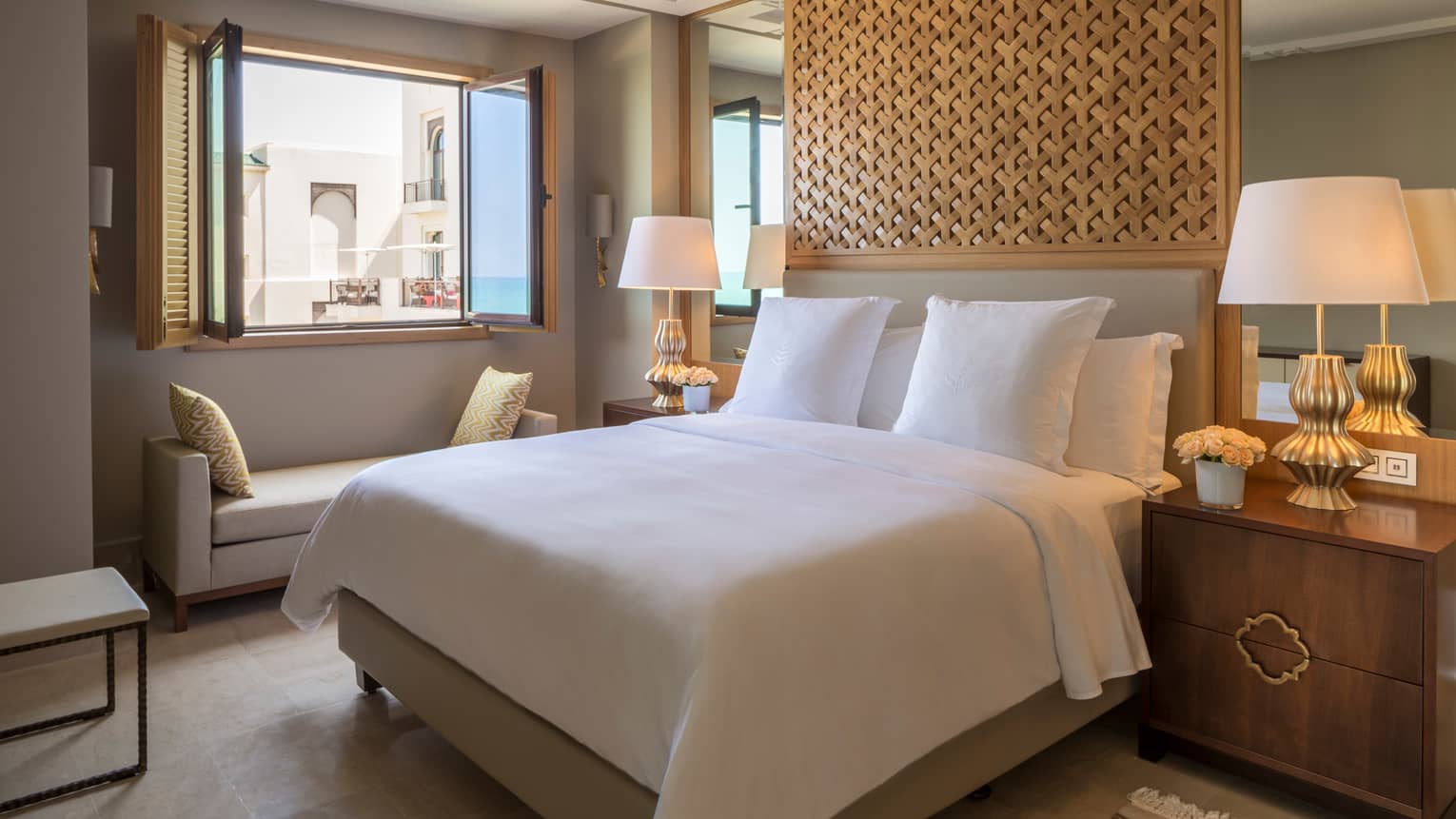 All-white bed with ceiling-high carved wooden headboard flanked by lamps, bench under window