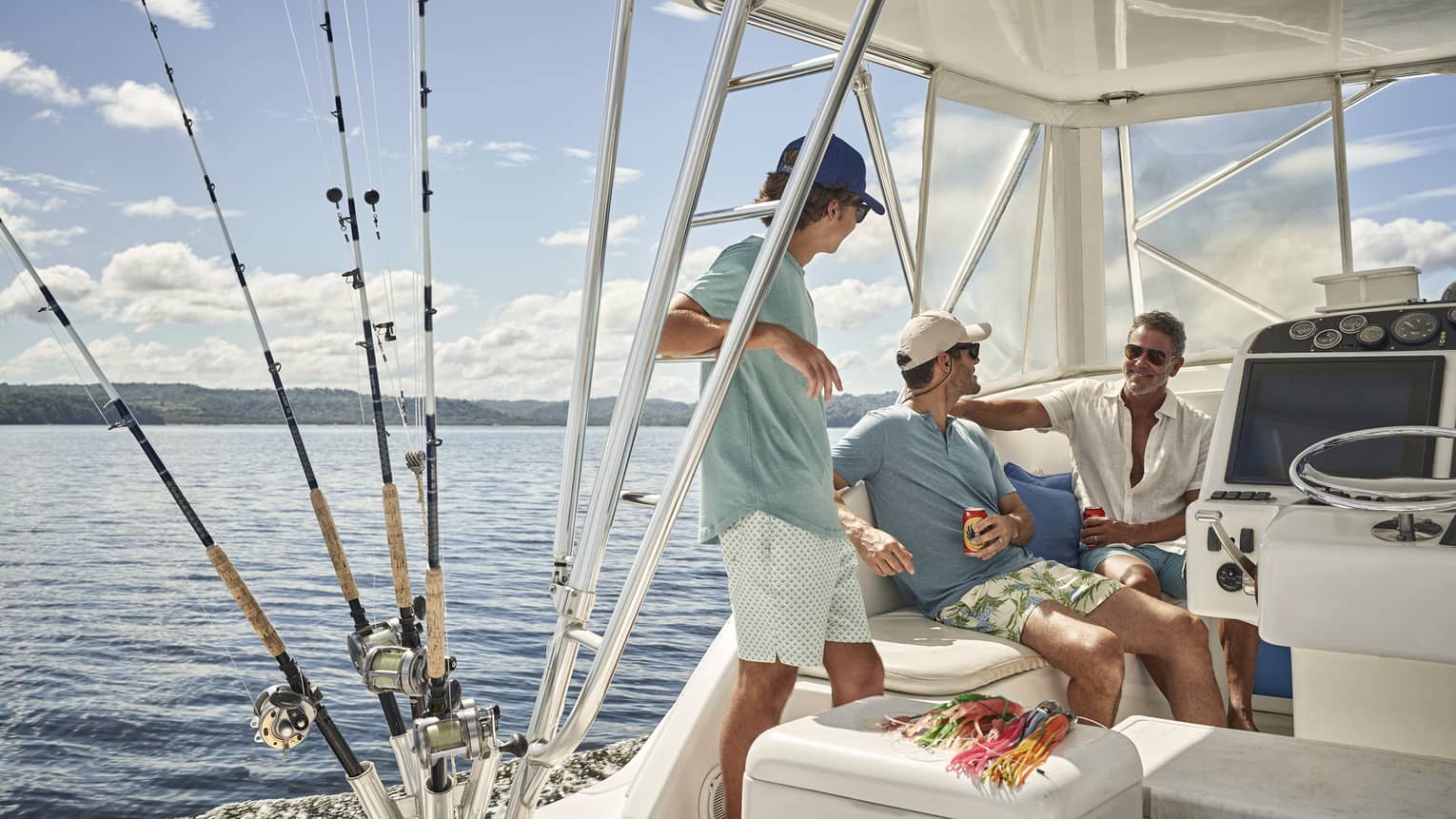 Aboard a fishing vessel on calm water, three people relax under the shaded cockpit, their fishing rods stored beside them. 