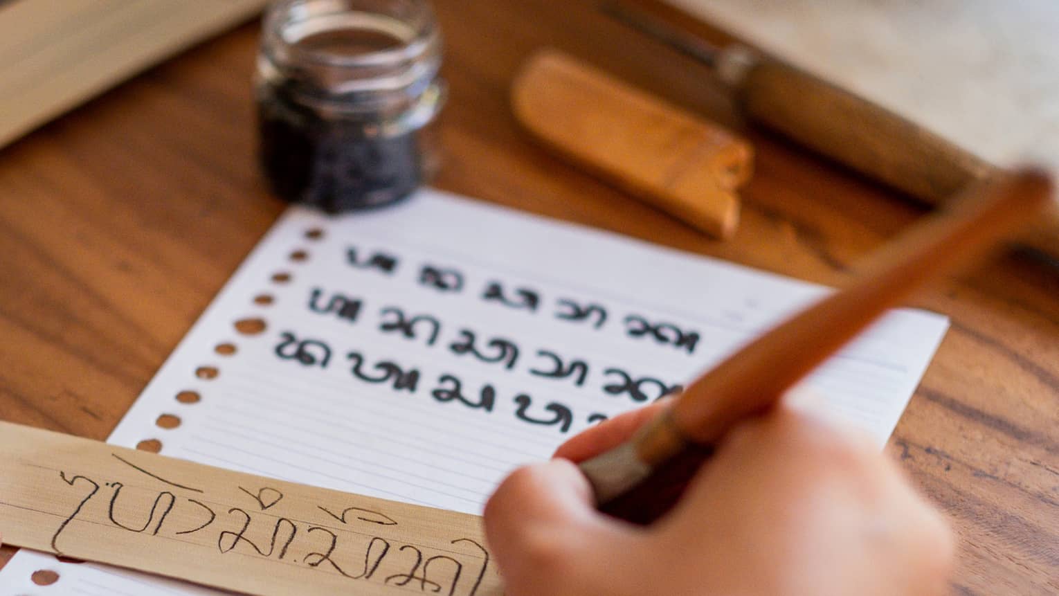 A hand holding a traditional wooden calligraphy pen copies Sanskrit text from a lined paper onto a dried palm leaf.