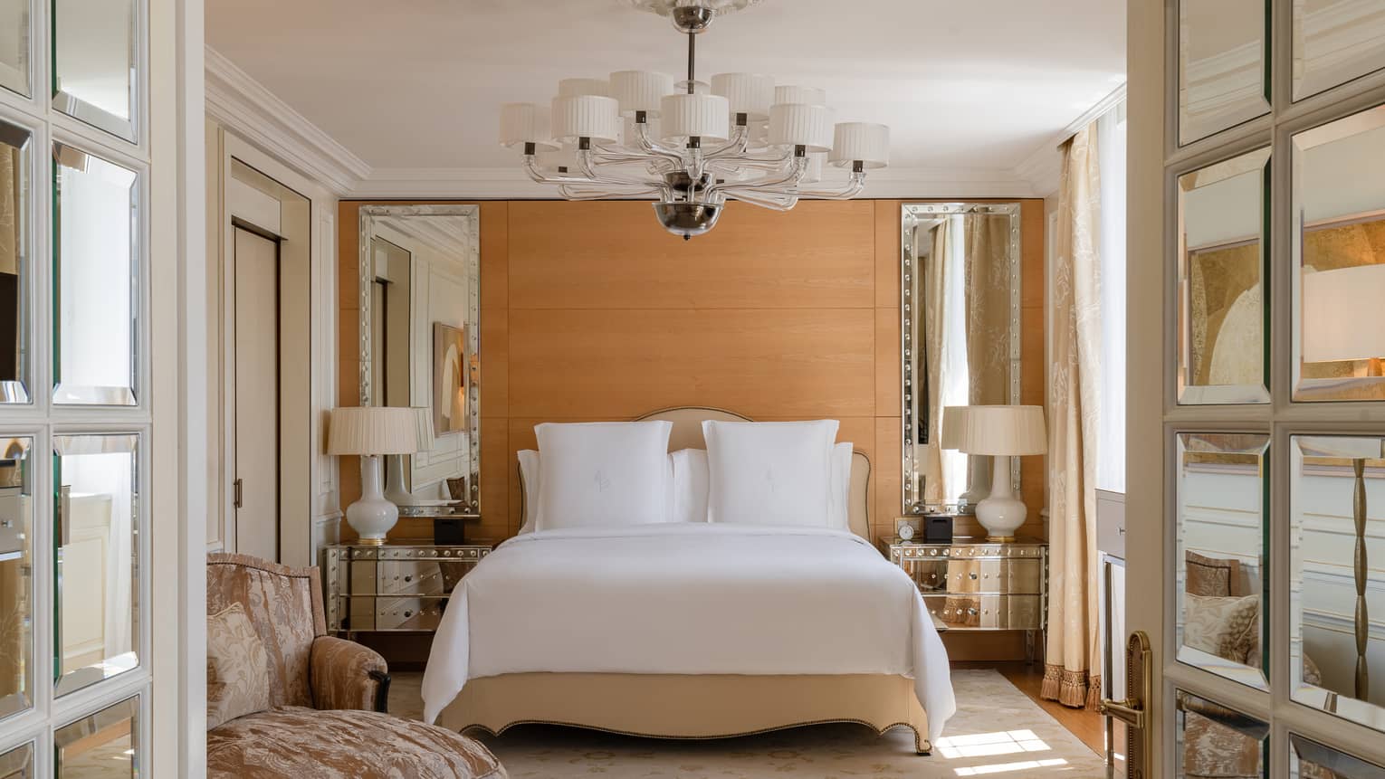 Luxury hotel bedroom with a king bed, mirrored bedside tables with lamps and a modern chandelier, complemented by soft beige accents and an elegant armchair