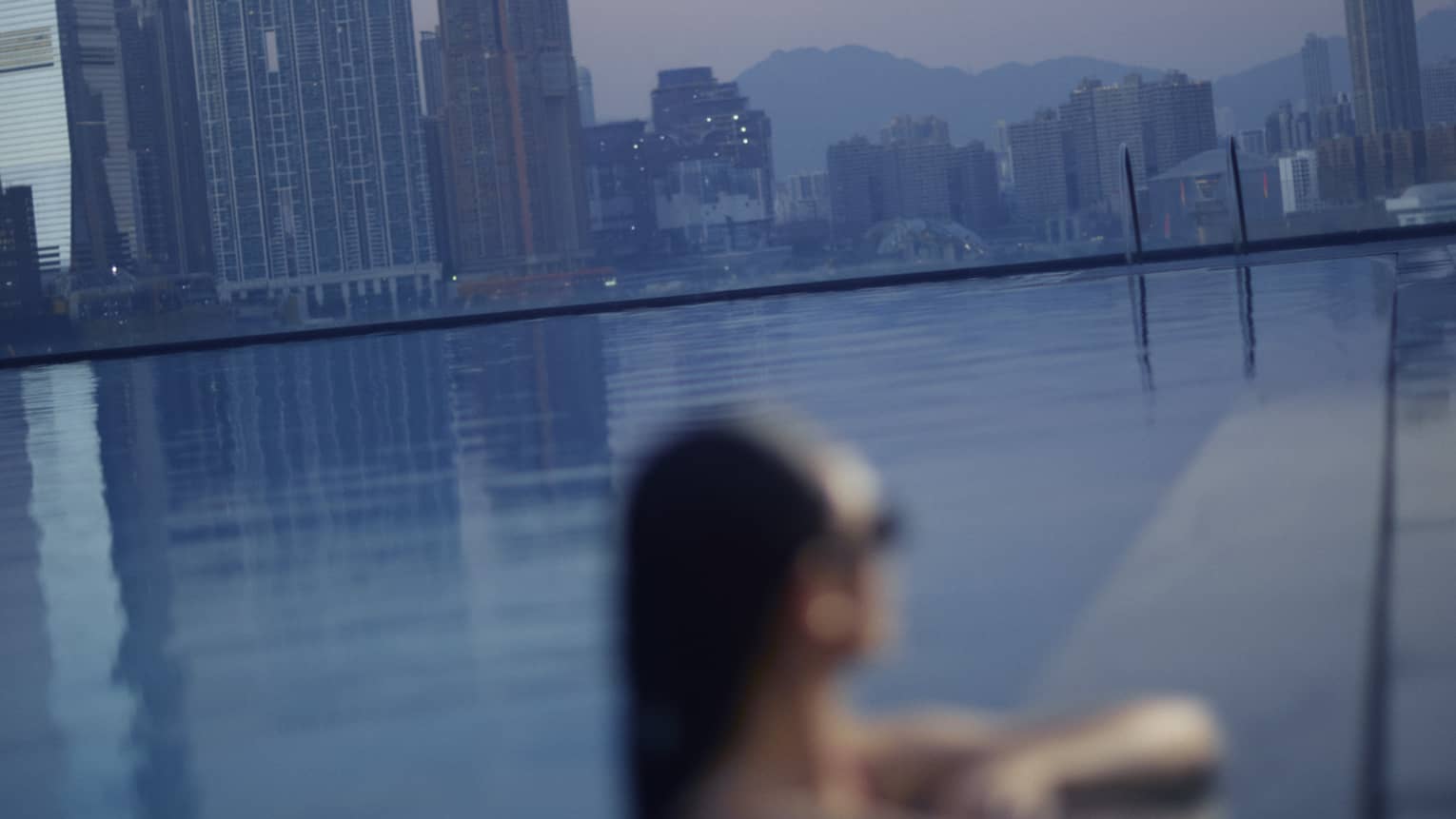 A woman in pool relaxes with elbows on pool deck, overlooking a city at dusk