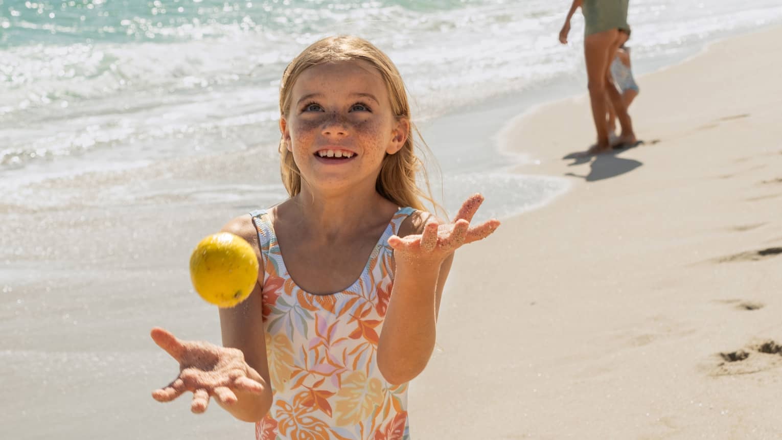 Child juggling lemons on a sunny beach with gentle ocean waves in the background, while another person walks along the shoreline in the distance.
