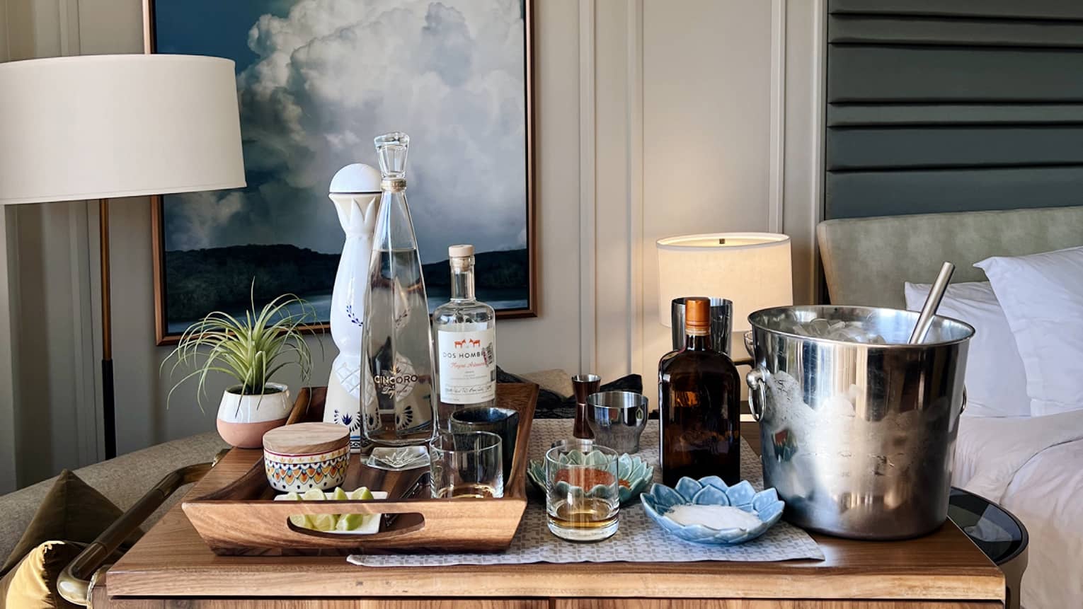 In a guest room, a wood cabinet is topped with clear bottles, glasses, a jigger, an ice bucket and dishes of salt and limes.