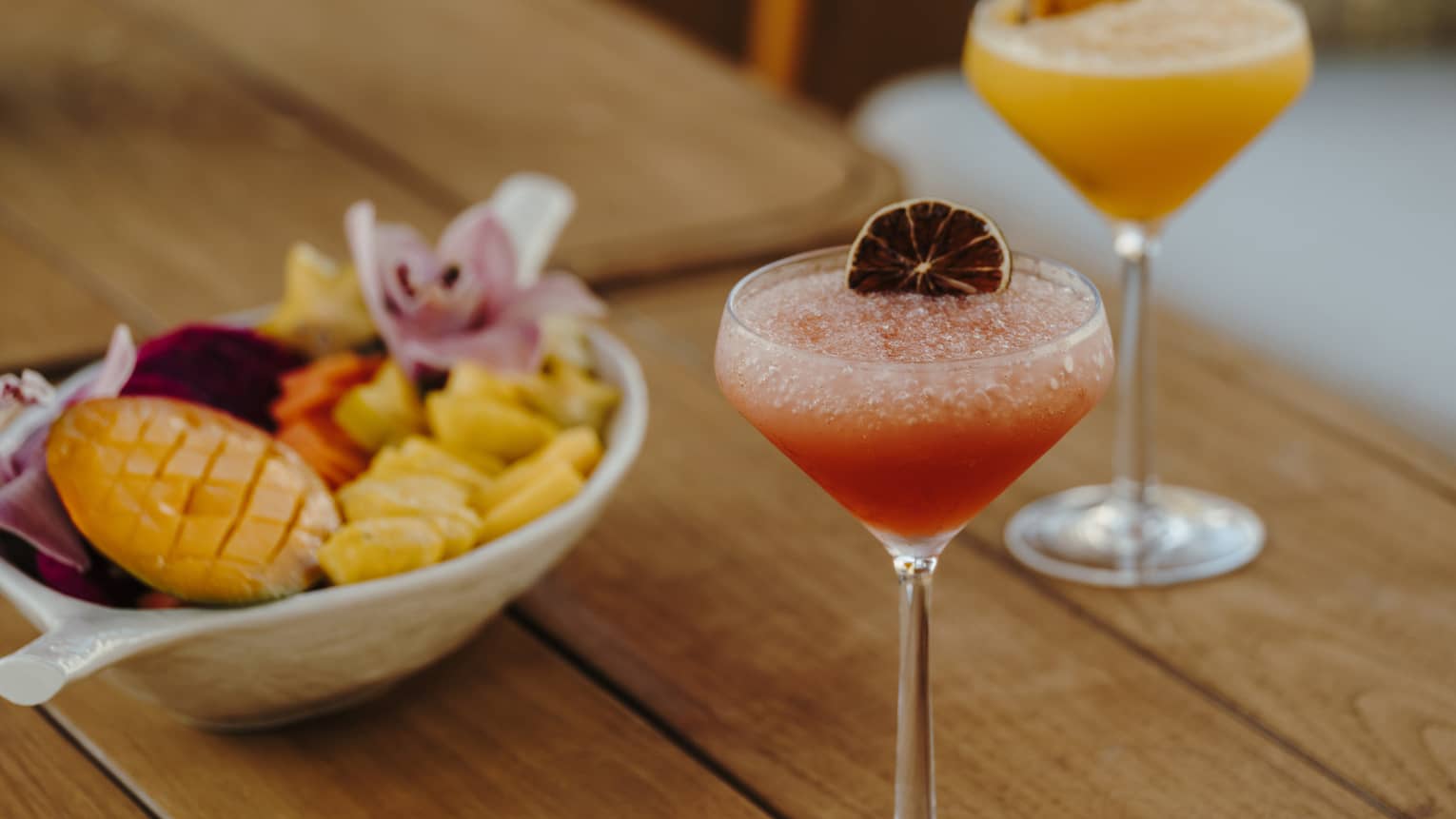 A wood table holds two stemmed glasses of pink and orange drinks with garnishes and a bowl of small-bite food items.