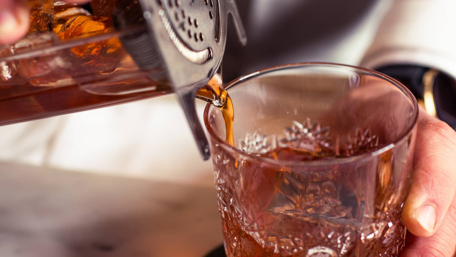 A cocktail shaker is tilted on an angle as dark amber liquid flows into an ornate cut-glass lowball cocktail tumbler.