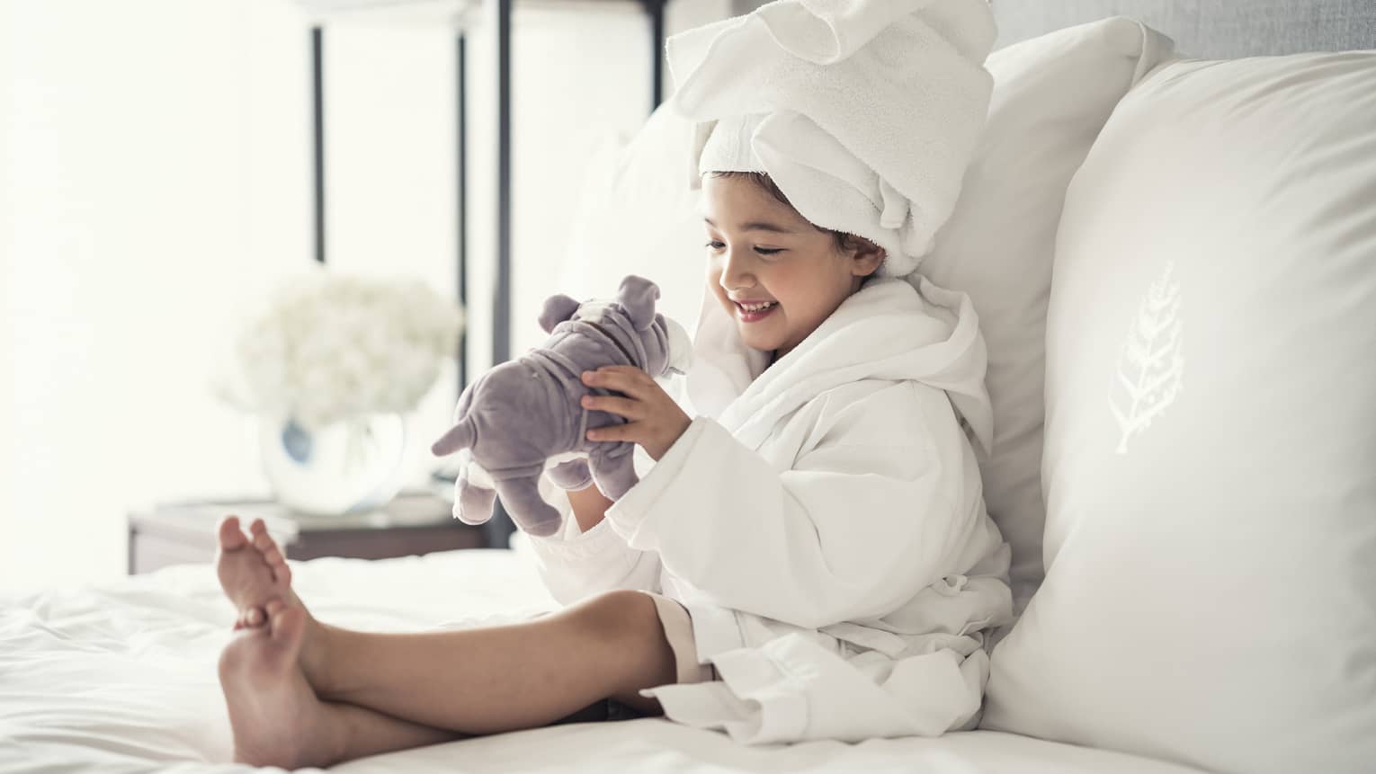 Young girl wearing a white bathrobe with towel wrap plays with plush dog toy on bed
