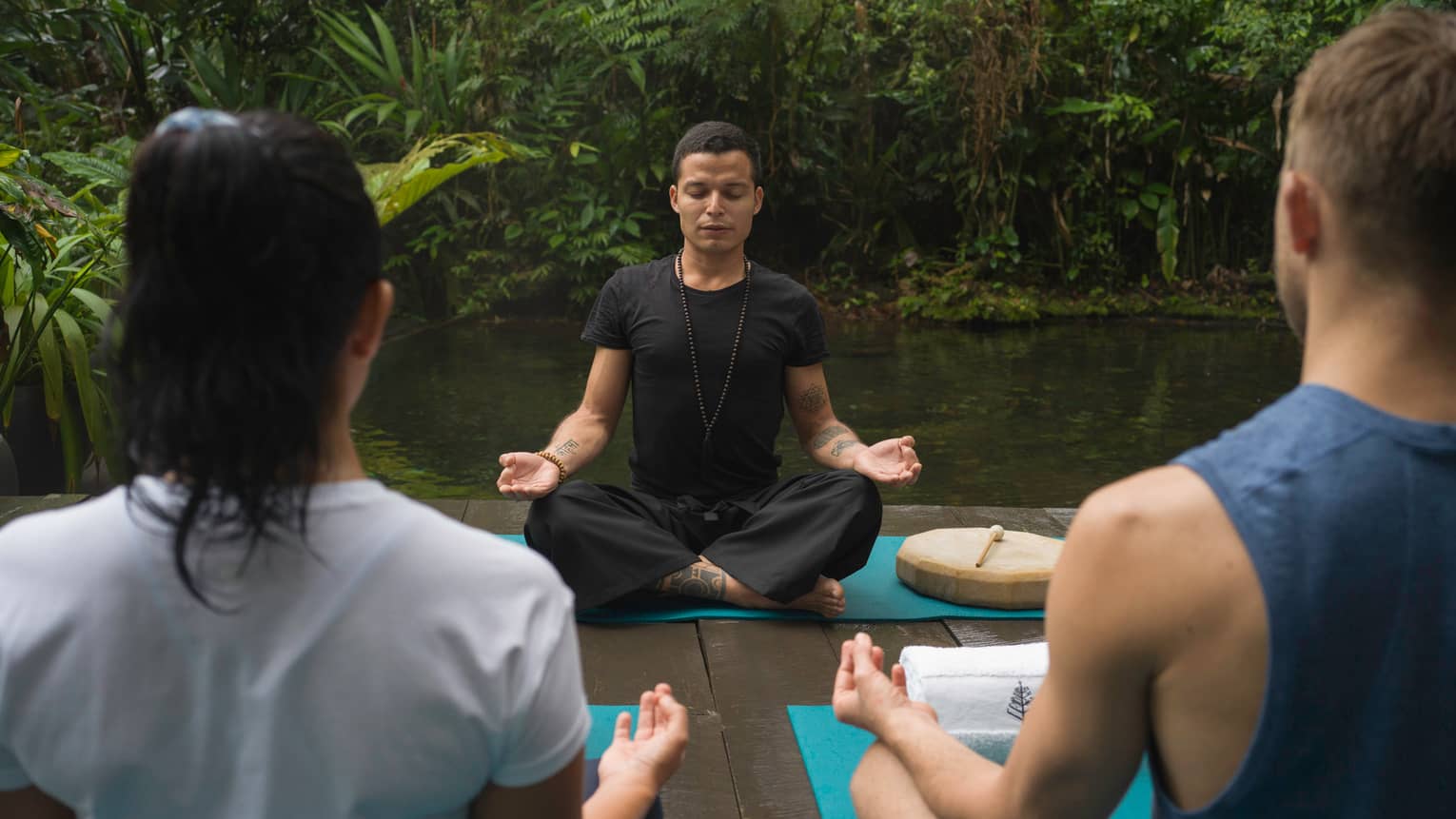Two people sit with their backs to the camera looking onto an instructor who is sitting cross-legged and with hands in the Gyan mudra position