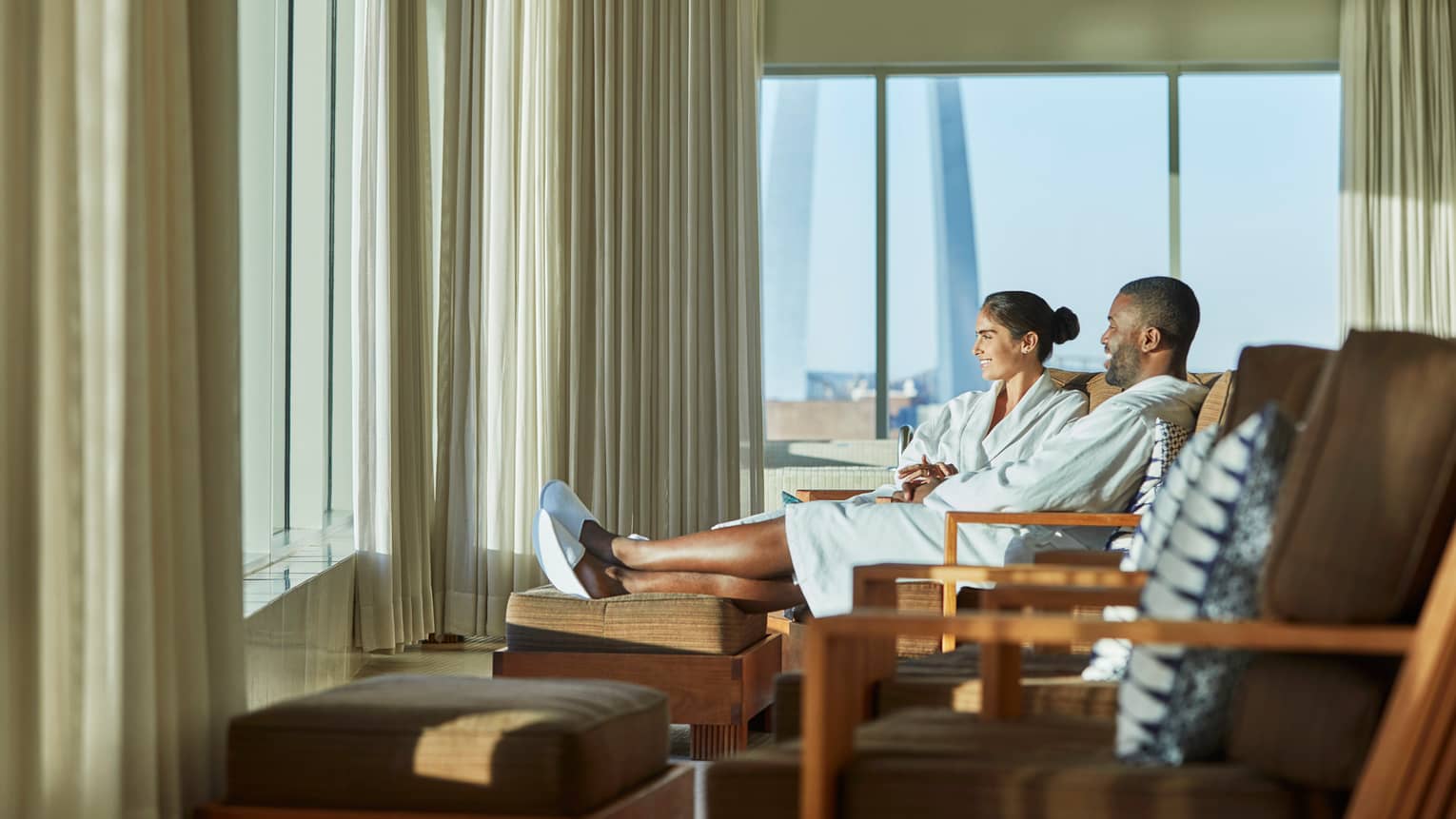 Smiling man and woman in bathrobes and slippers sit on lounge chairs near sunny window