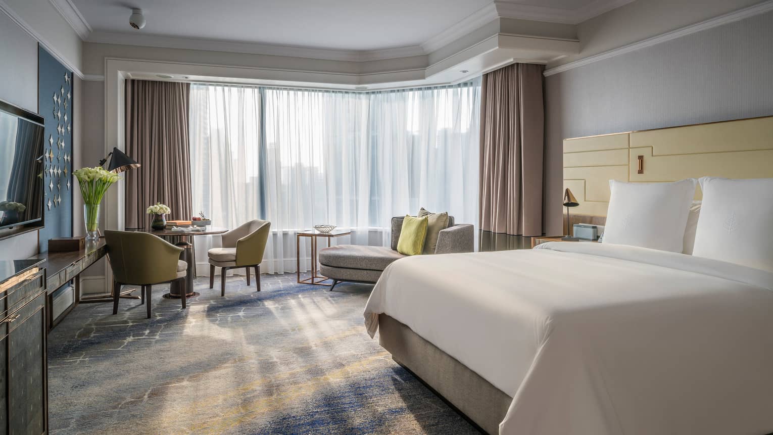 Sunny Deluxe Room bed with white linens, small dining table and chaise lounge, white curtains