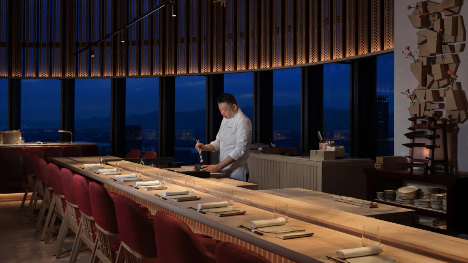 Chef preparing sushi at Sushi L’Abysse Osaka Yannick Alléno, with a scenic night view and elegant counter seating.