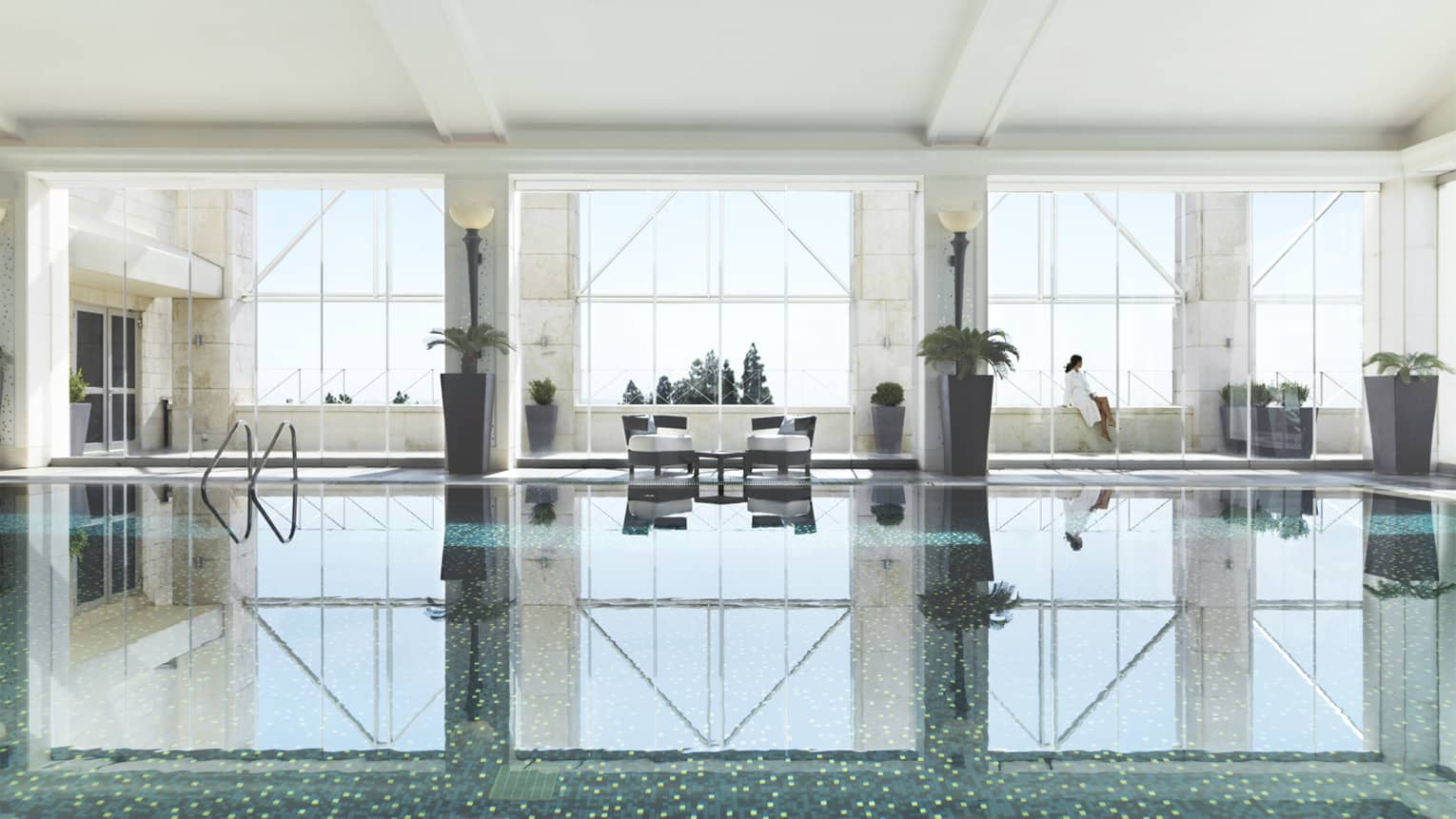 Indoor swimming pool reflecting three tall windows, woman in white bathrobe sits on ledge