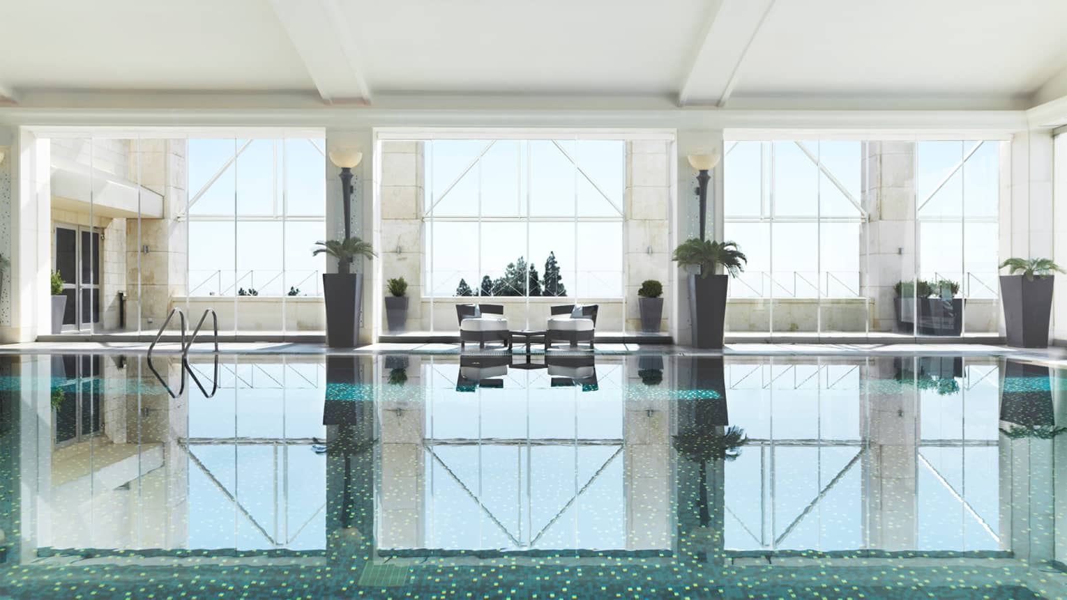 Indoor swimming pool reflecting light of three tall windows, white walls and pool chairs 